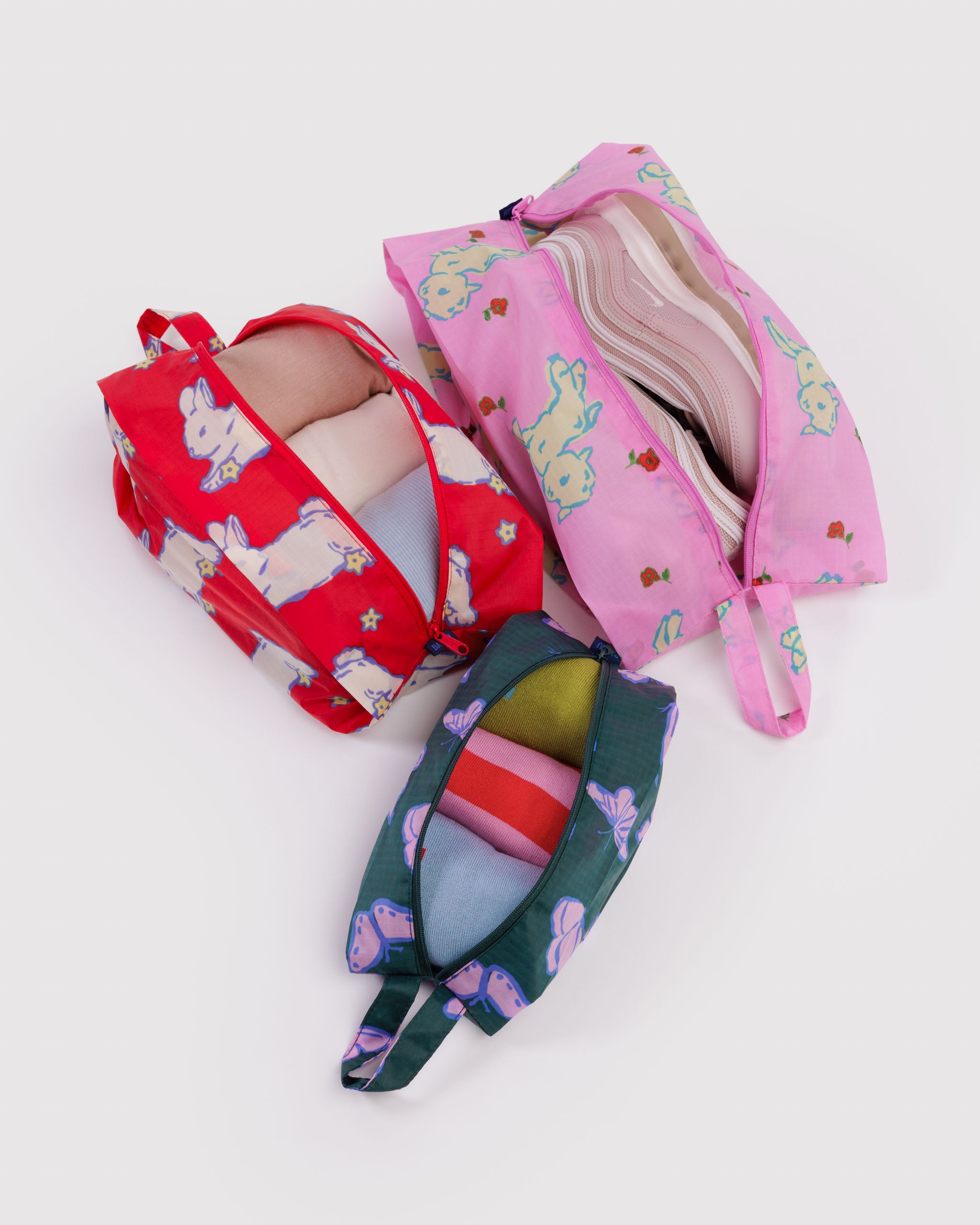 Three colourful pouches with different animal designs with zip top open sitting flat on bottom and expanded with clothing and shoes items inside.