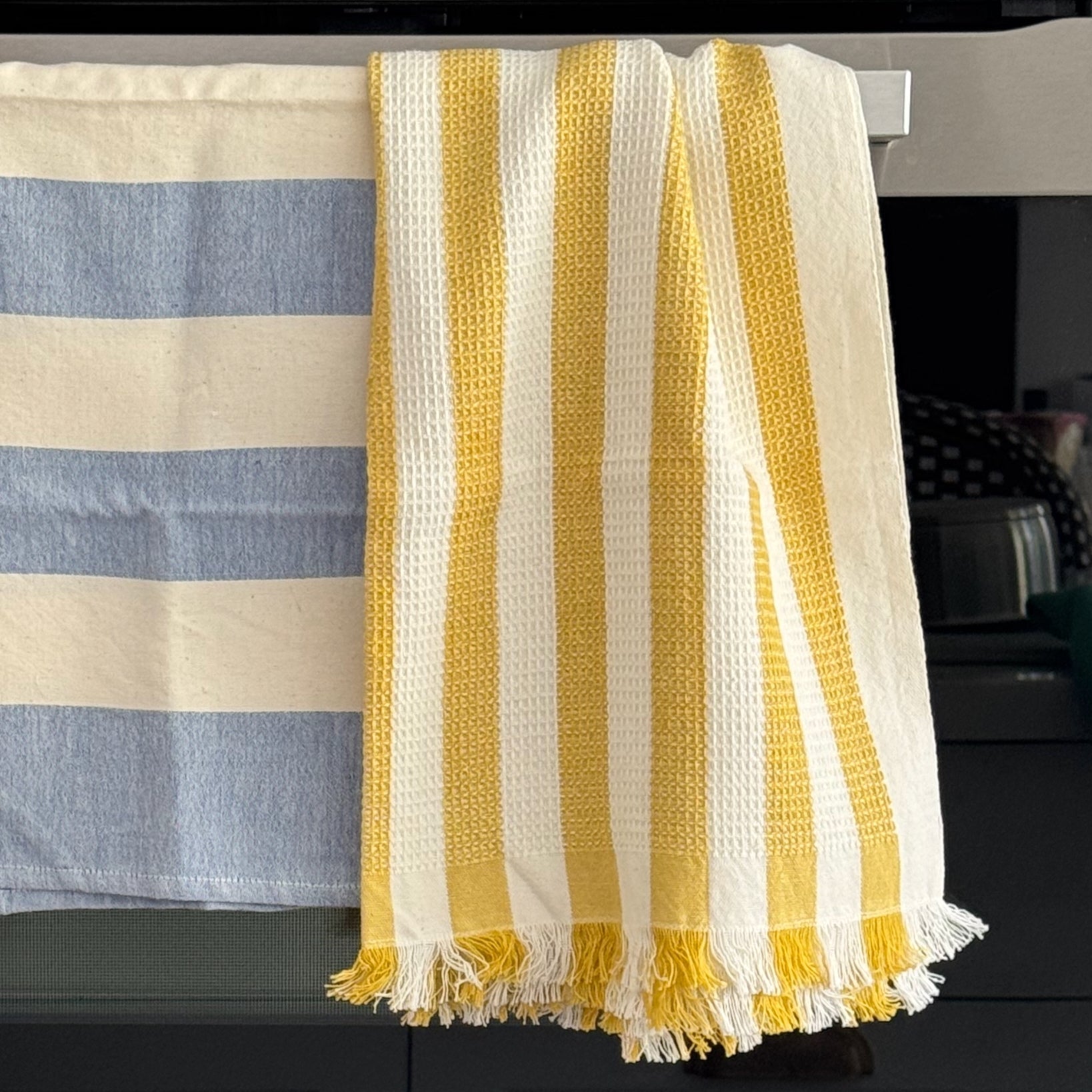Blue and white wide striped tea towel and a yellow and white waffle hand towel with fringe ends draped over oven handle.