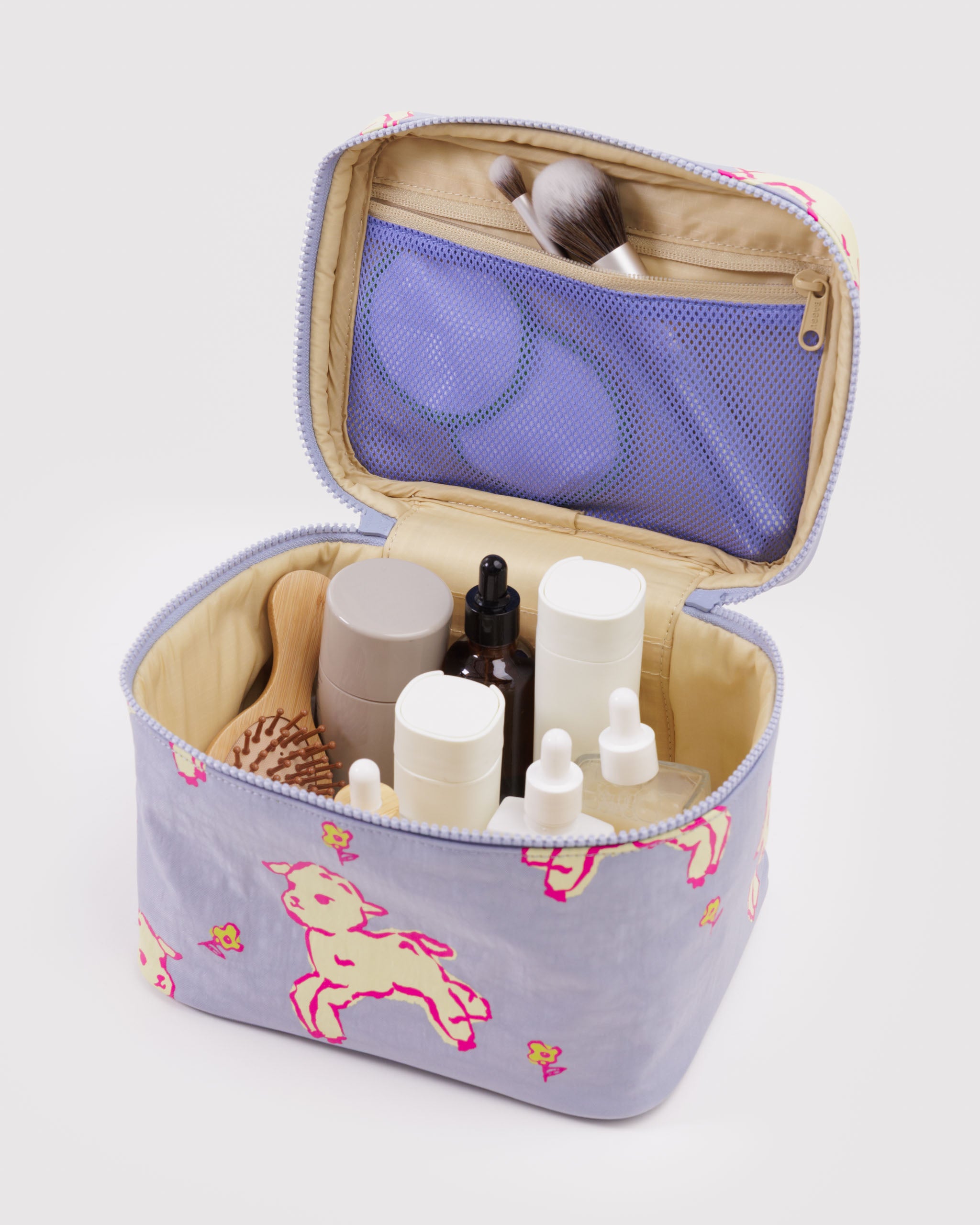 Open blue cosmetic bag with white lambs and yellow flowers pattern containing various skincare products and makeup brushes in zip mesh pouch on lid.