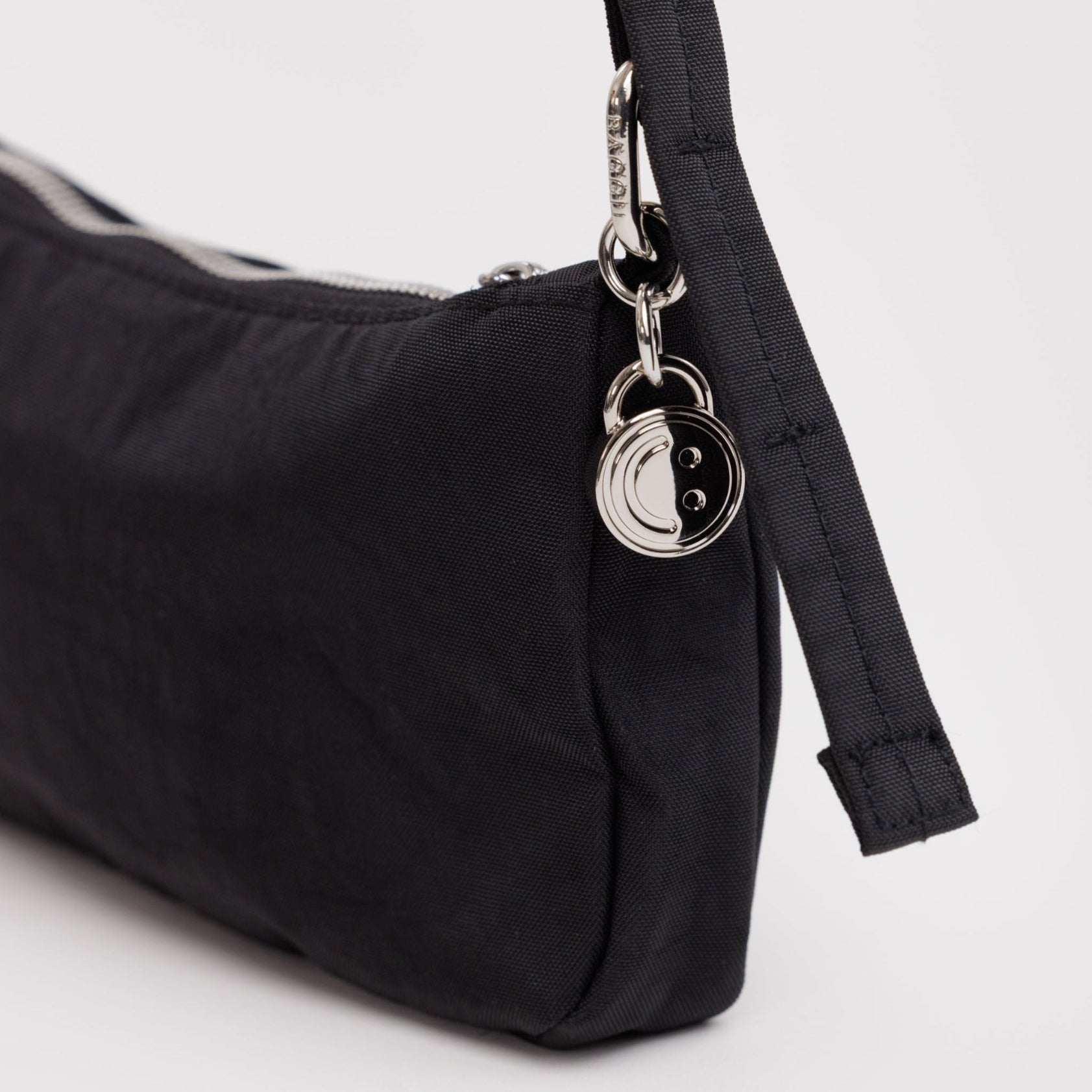Small nylon black print handbag with an adjustable strap and silver hardware clasp, smiley face charm and zip.