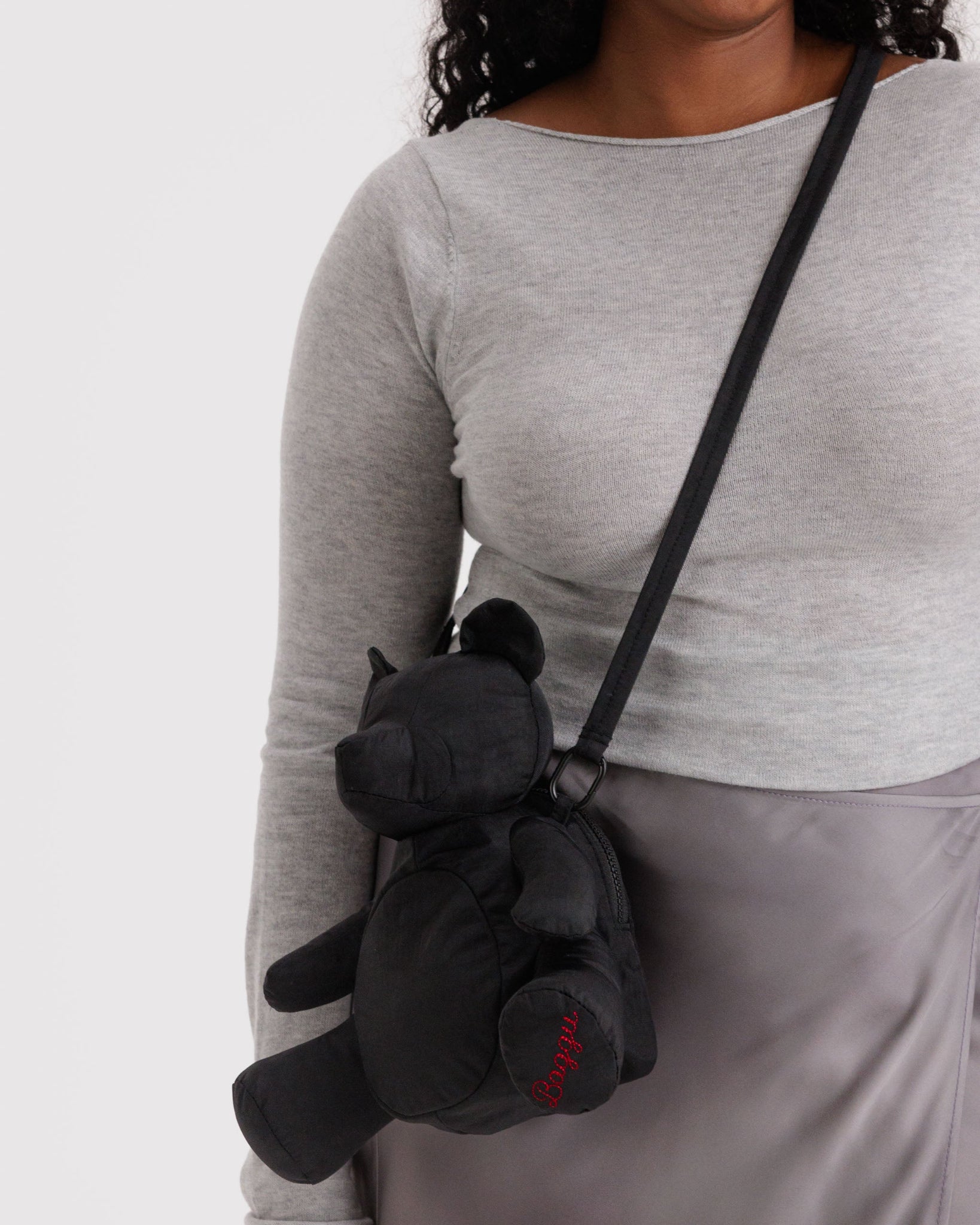 Person wearing a black teddy bear-shaped bag with an adjustable crossbody strap and red BAGGU embroidery on foot.