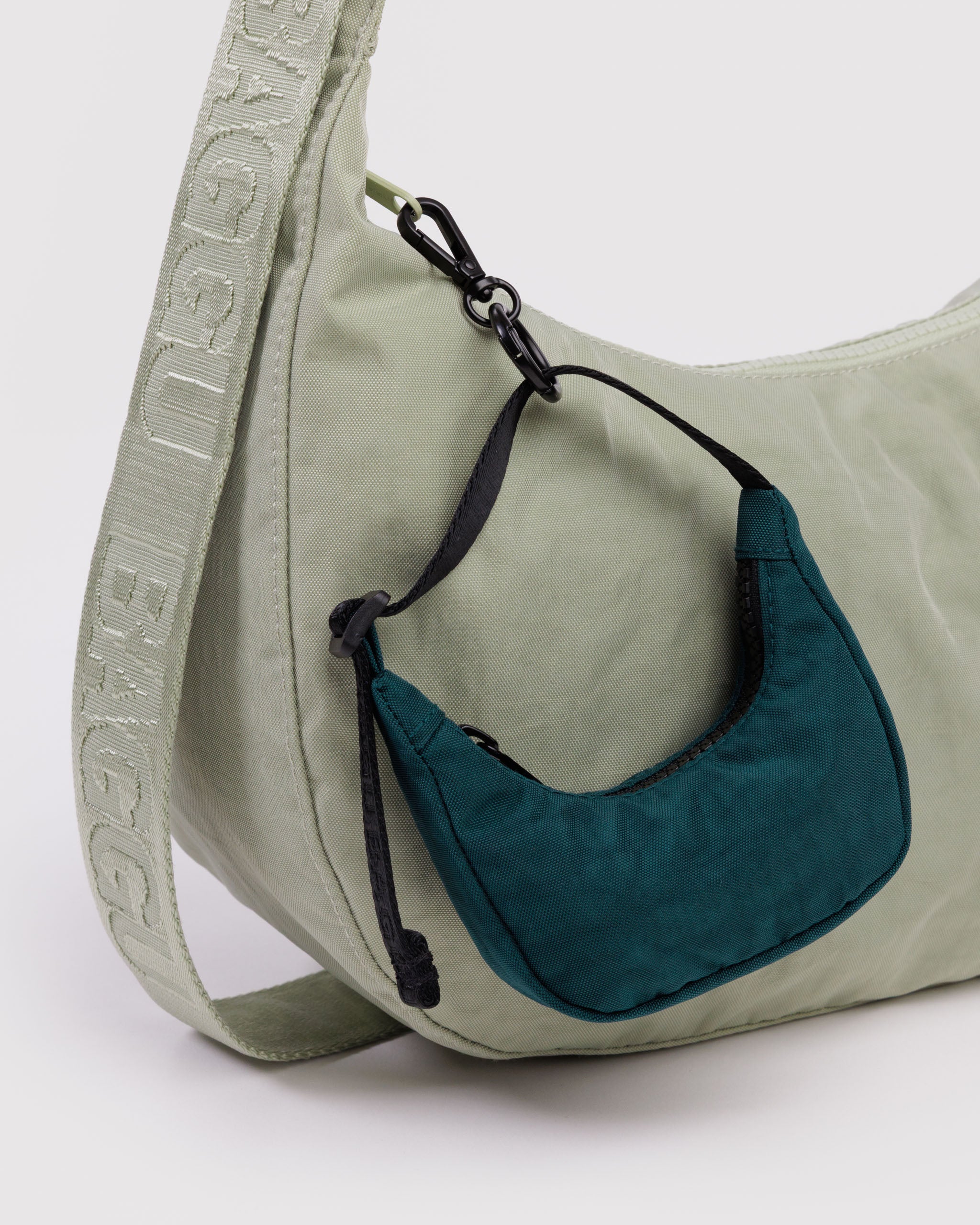 Sage coloured shoulder bag with a green crescent shaped bag charm attached to zipper with the black metal clip.