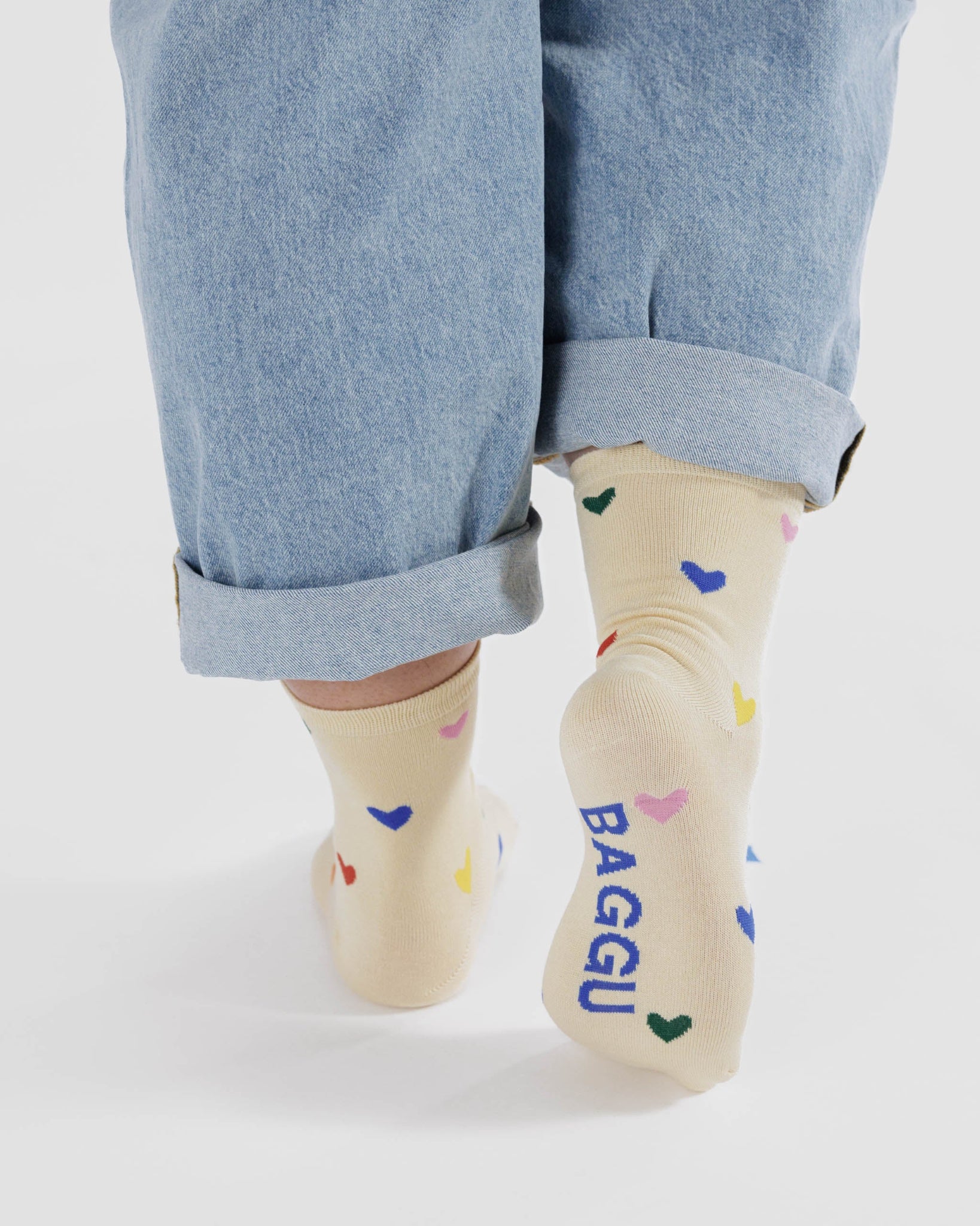 Person wearing cream socks with colourful heart patterns and 'BAGGU' branding on sole worn with rolled-up blue jeans.