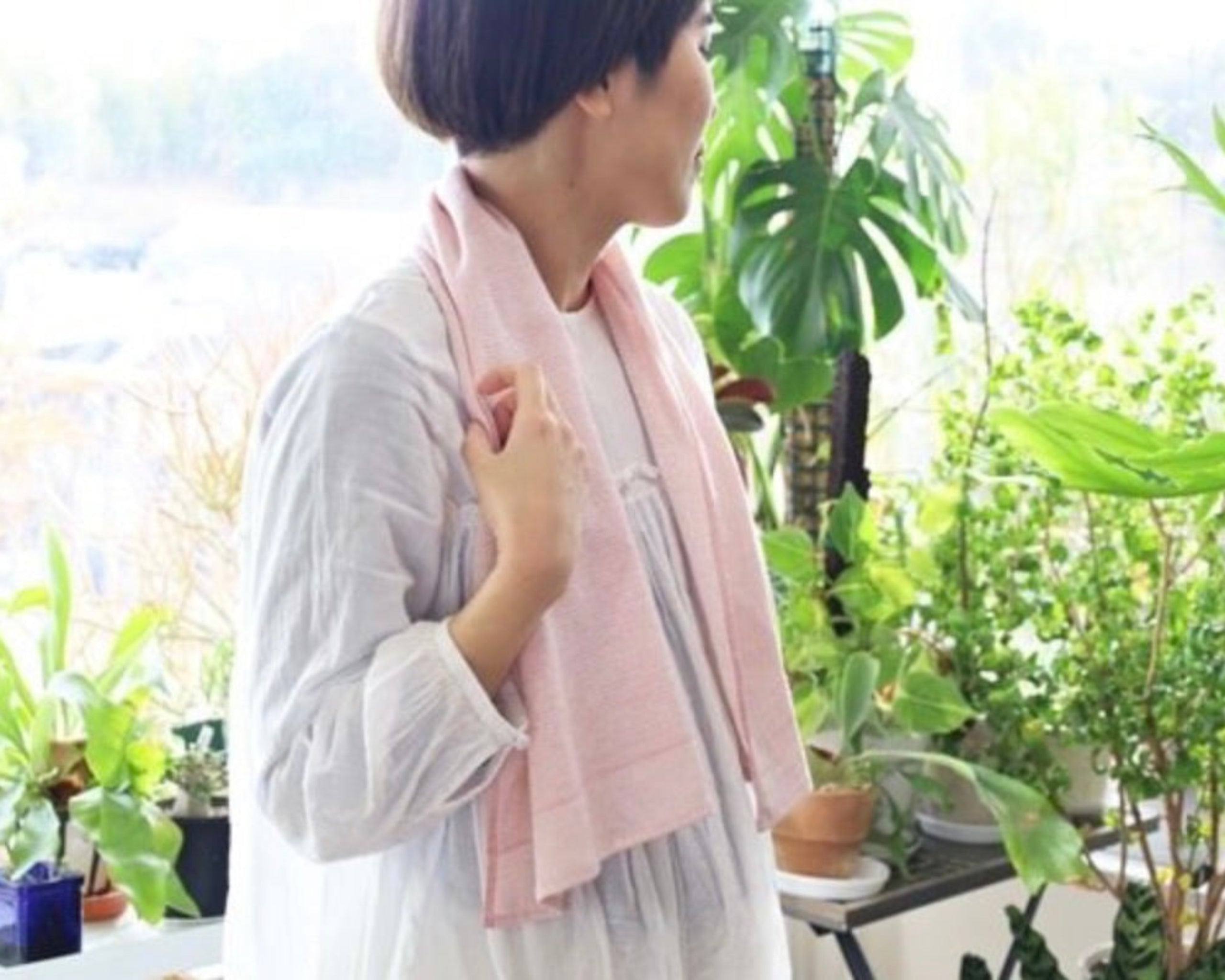 Person wearing a light pink towel around neck like a scarf in a room with plants.