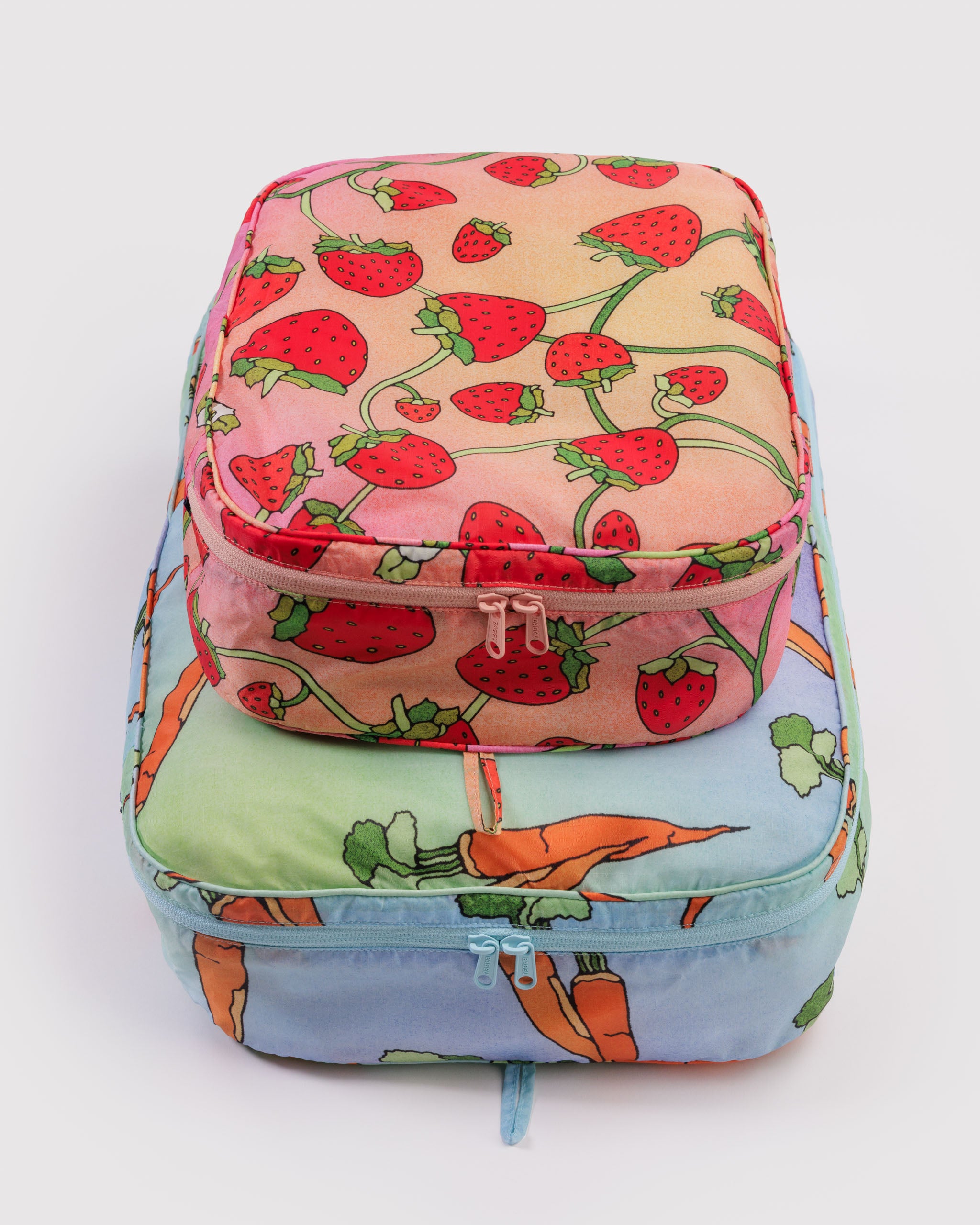 Two different sized colourful cubes with strawberry and carrot patterns and all around zippers and a hanging loop filled with items.