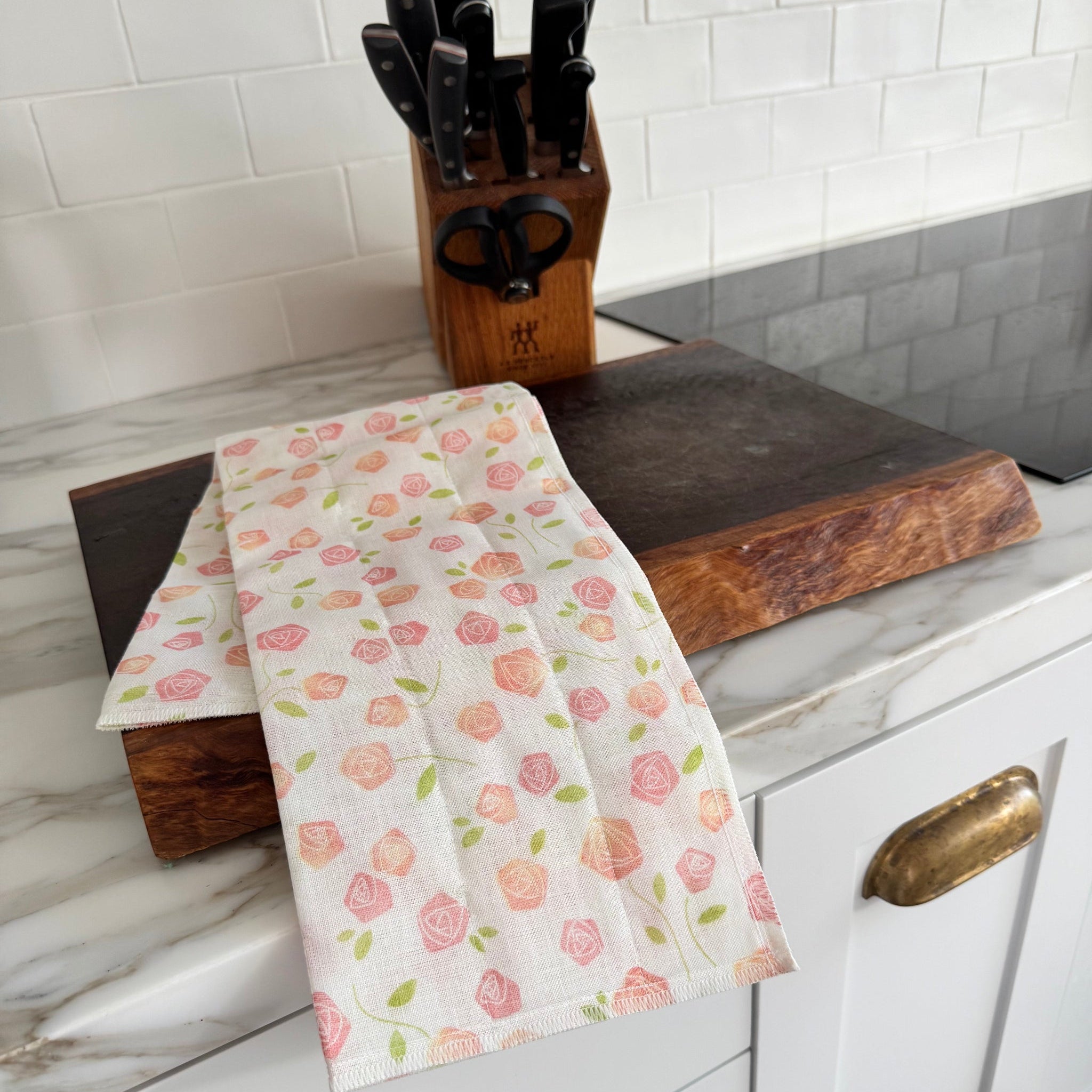 Floral rose tea towel with cream background draped over a wooden cutting board near a wooden knife block on a kitchen counter.
