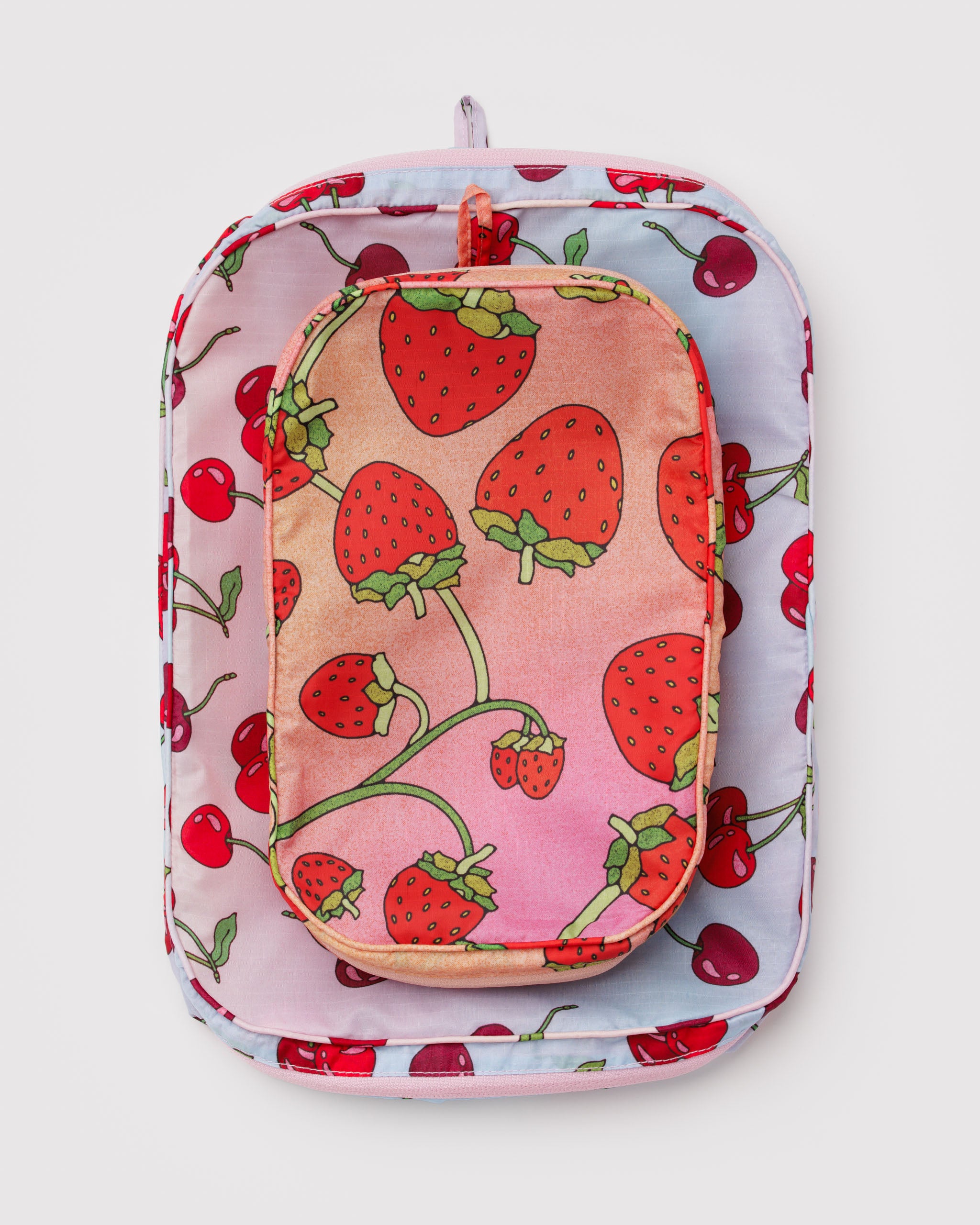 A stacked set of 2, a large and a medium size, packing cube with strawberry on ombre pink and orange fabric and a cherry pattern on ombre light blue and pink fabric with all around zippers and a hanging loop.