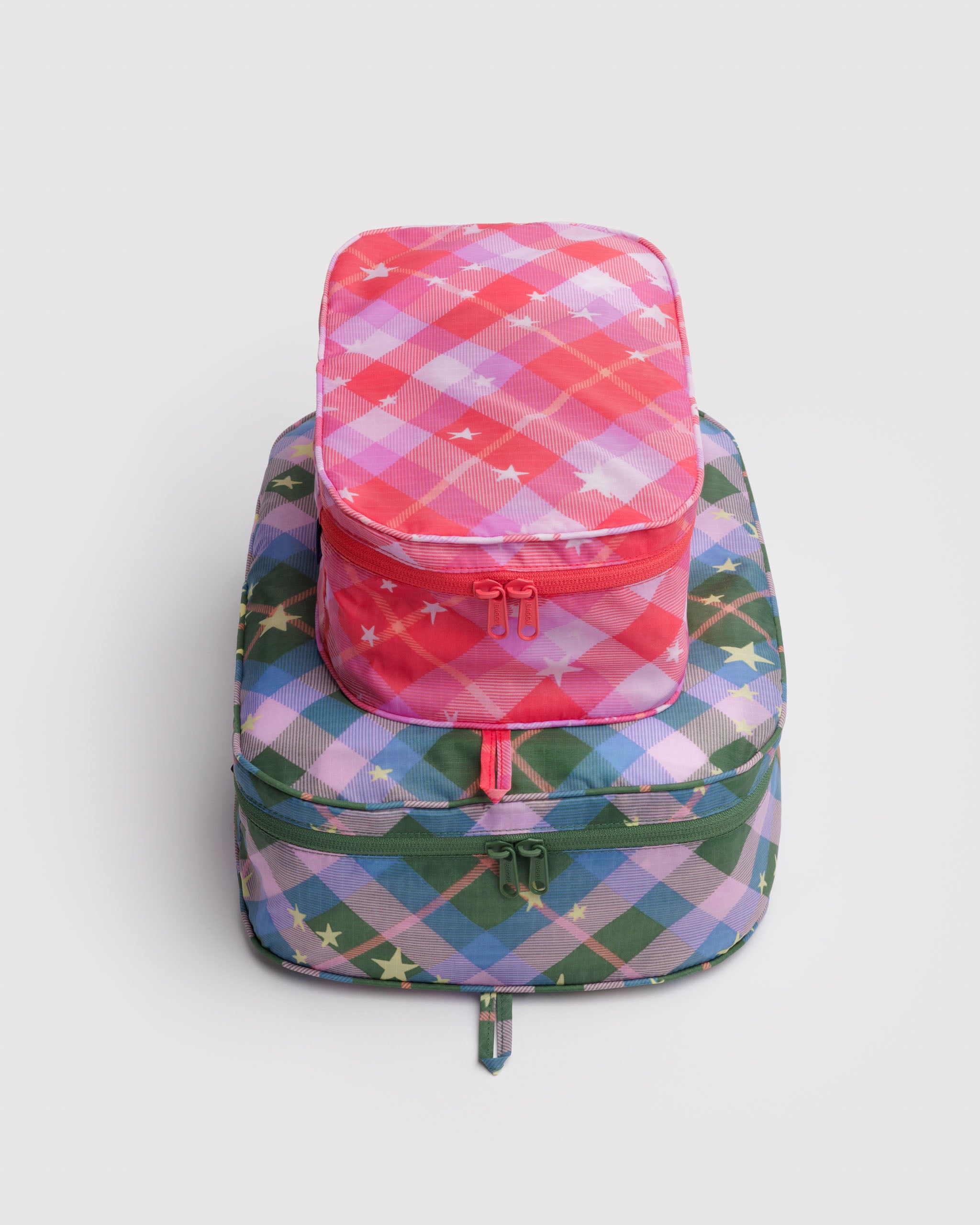 Stack of two packed, star plaid patterned packing cubes with tonal zips and hanging loops, in a pink and a green.