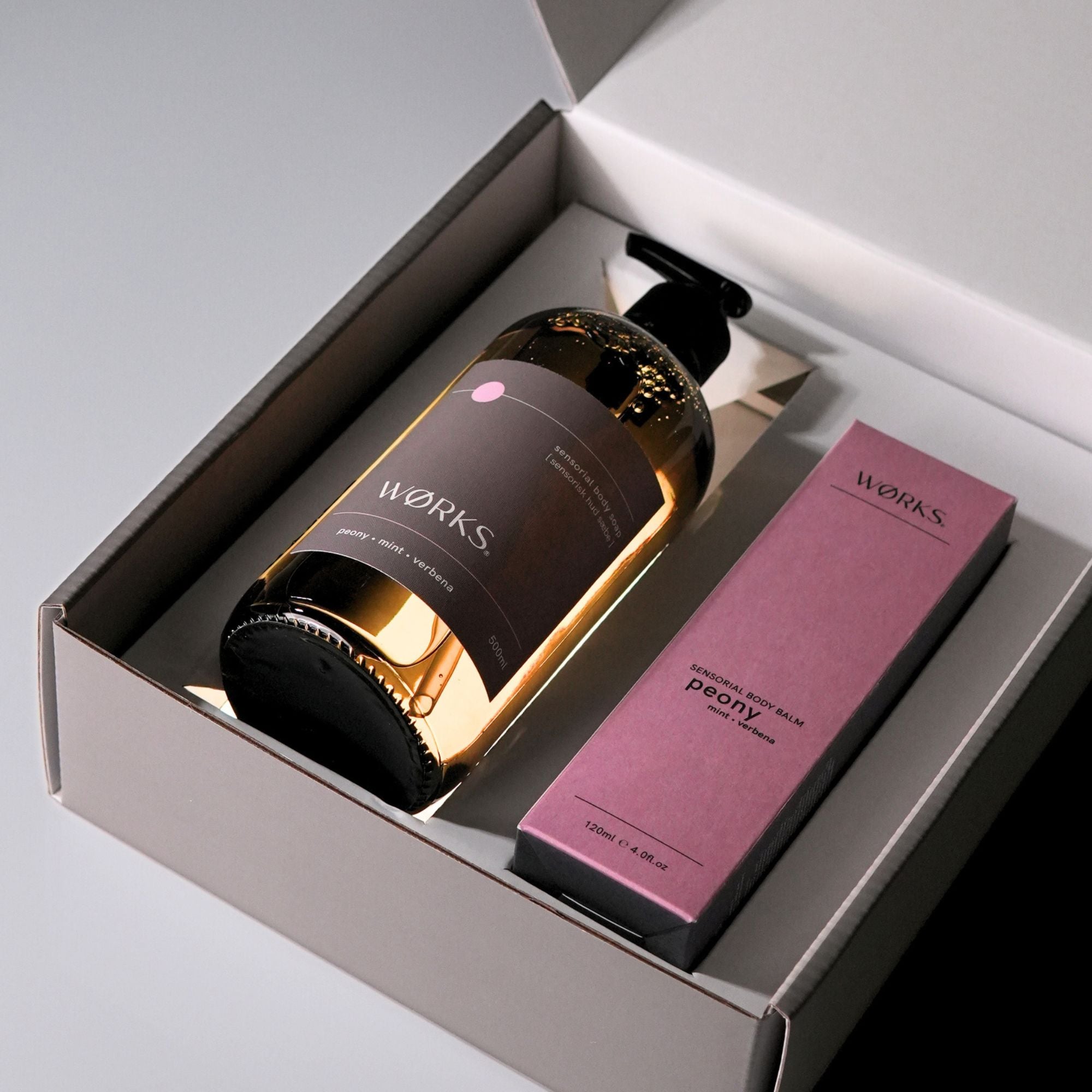 A brown, glass pump bottle and a pink box of 'WORKS' products in a grey gift box with lid open.