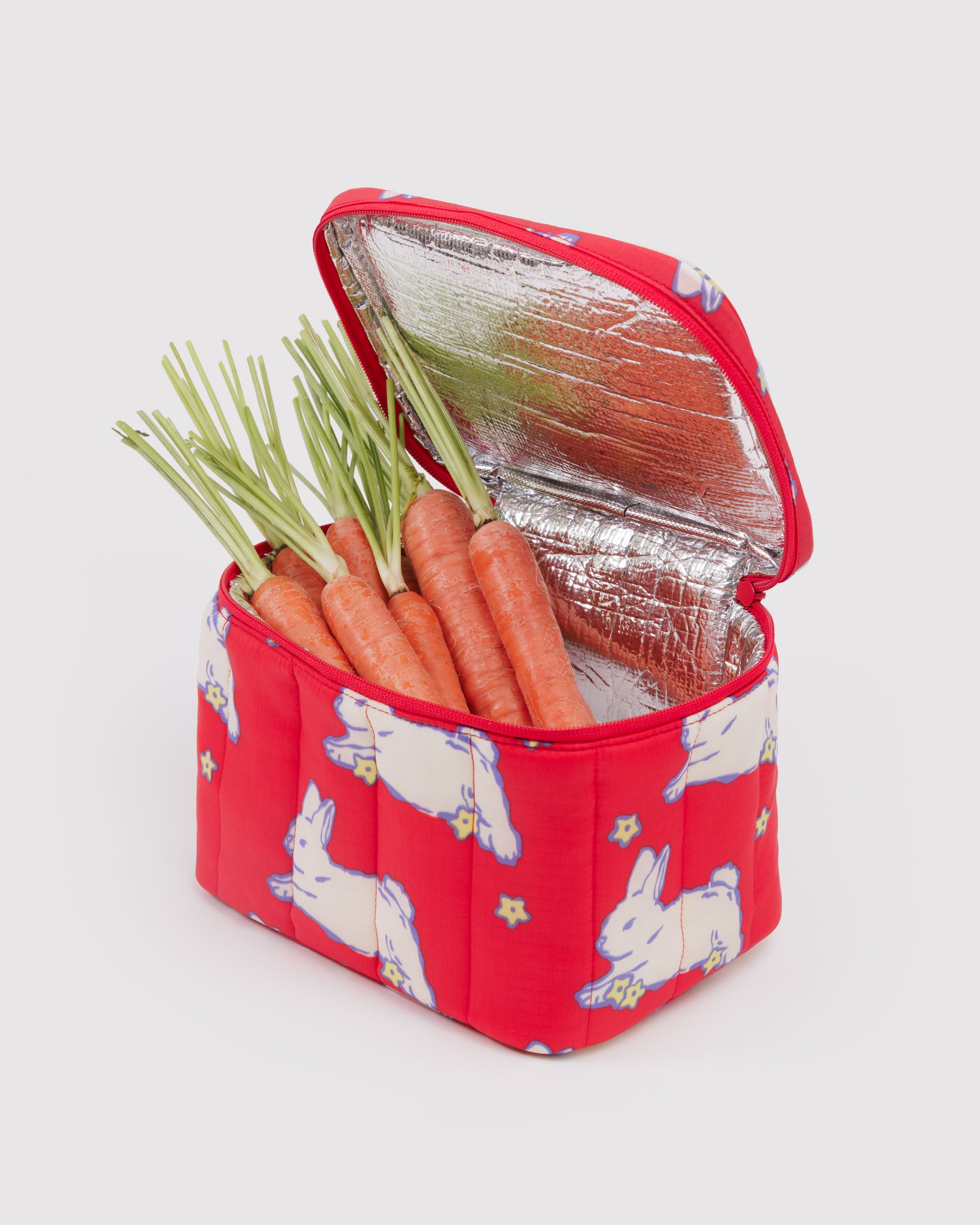 Red insulated lunch bag with white rabbit pattern with zip top open and containing a bunch of carrots with green tops.