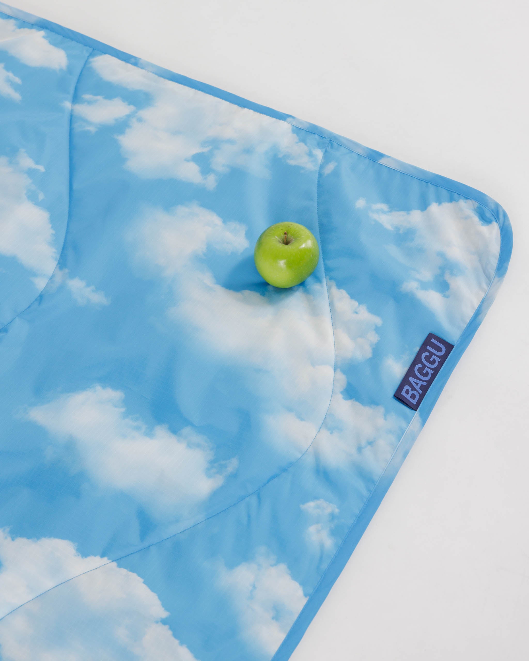 baggu-picnic-blanket-in-clouds-lying-flat-with-a-green-apple-on-top-corner