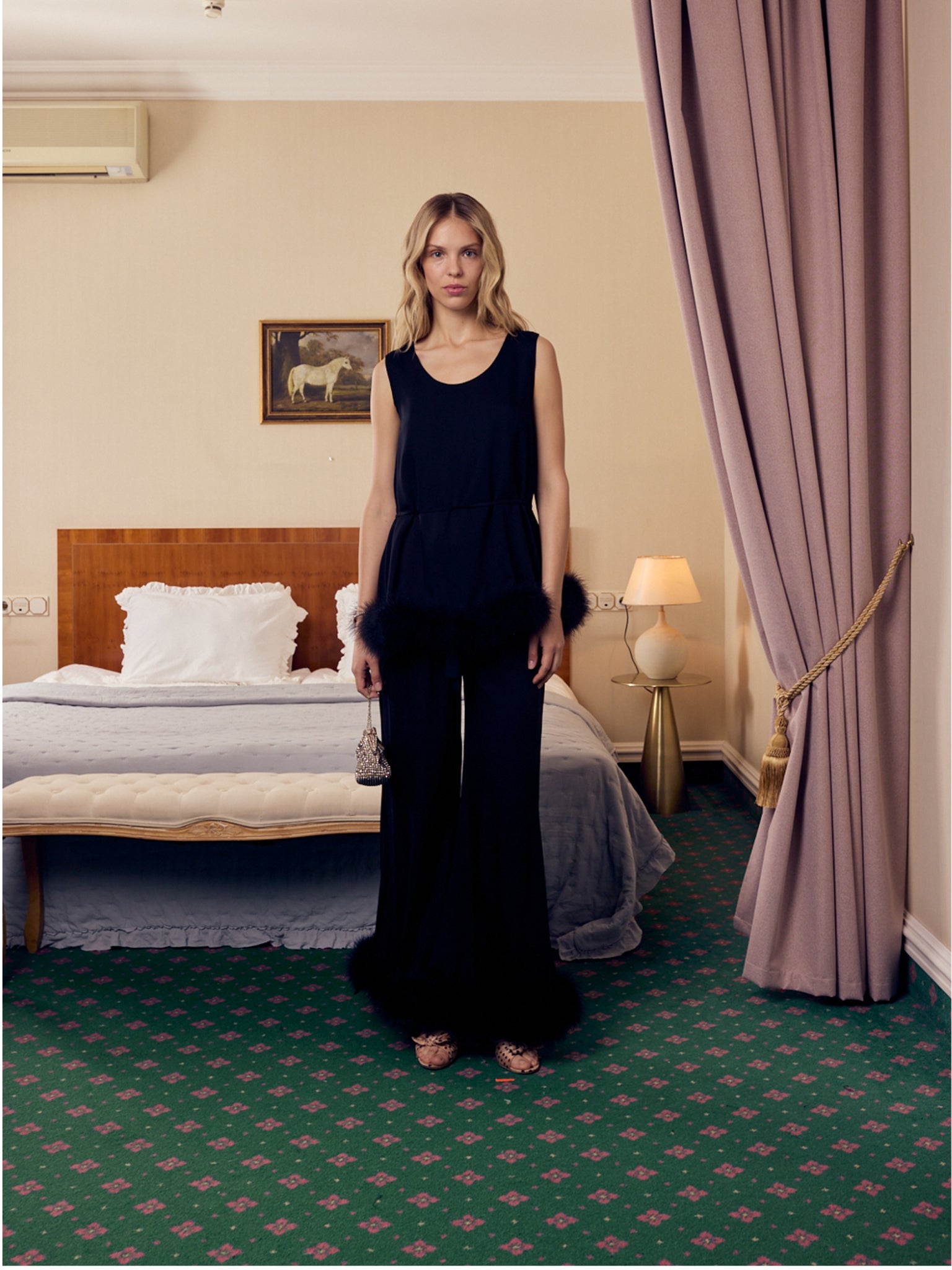 Woman in a black long pants and sleeveless top with feather trim outfit standing in a bedroom.