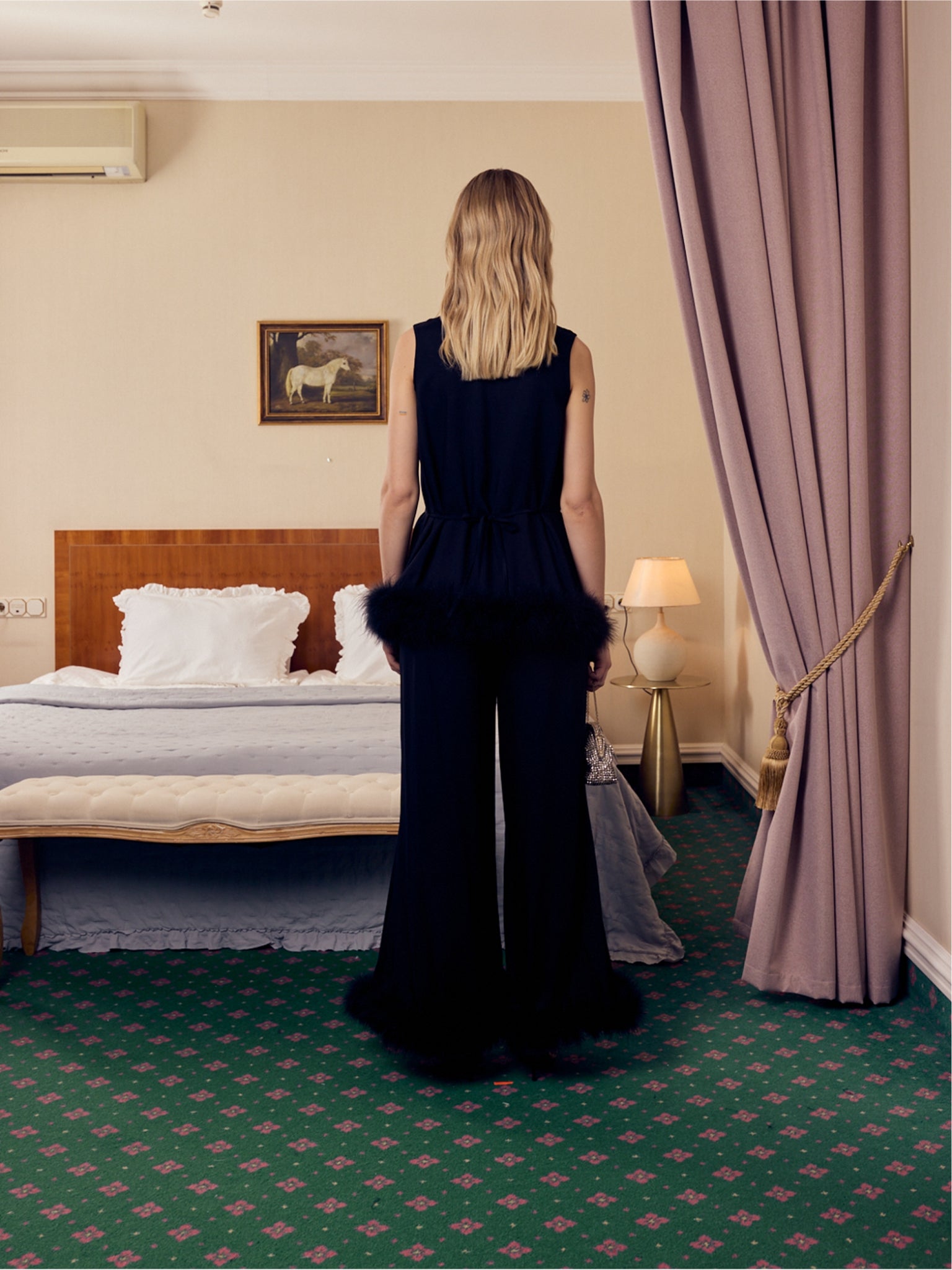Woman in a black top and long pants with black feather trim standing in a hotel room with a bed and lamp.