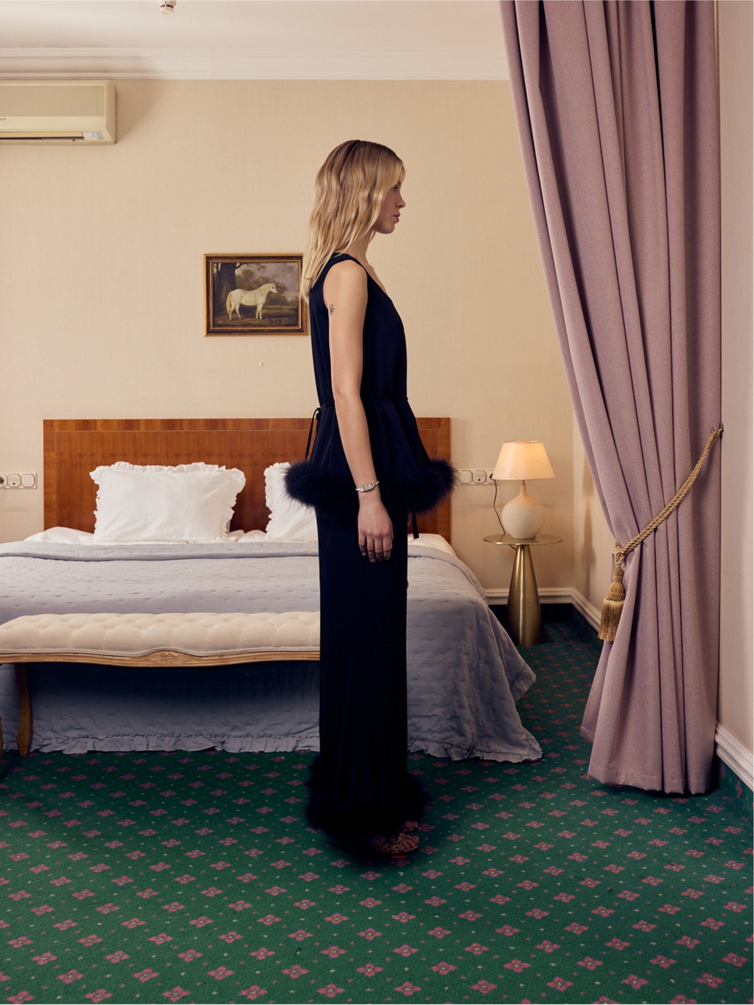 Side view of woman in a black pants and top with feather trim outfit standing in a bedroom with a bed and lamp in the background.