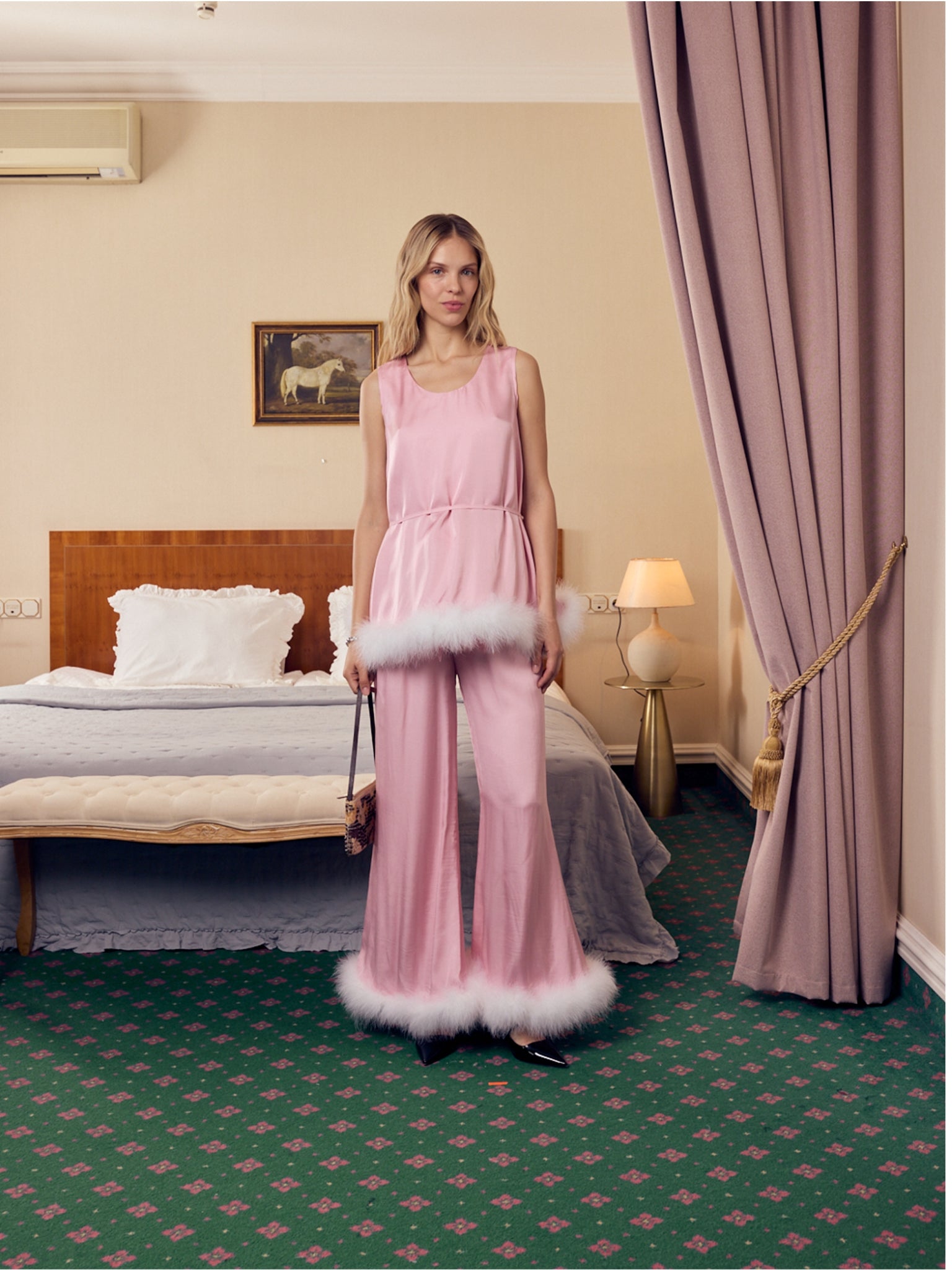 Woman in a pink sleeveless top and pants with white feather trim standing in a bedroom.