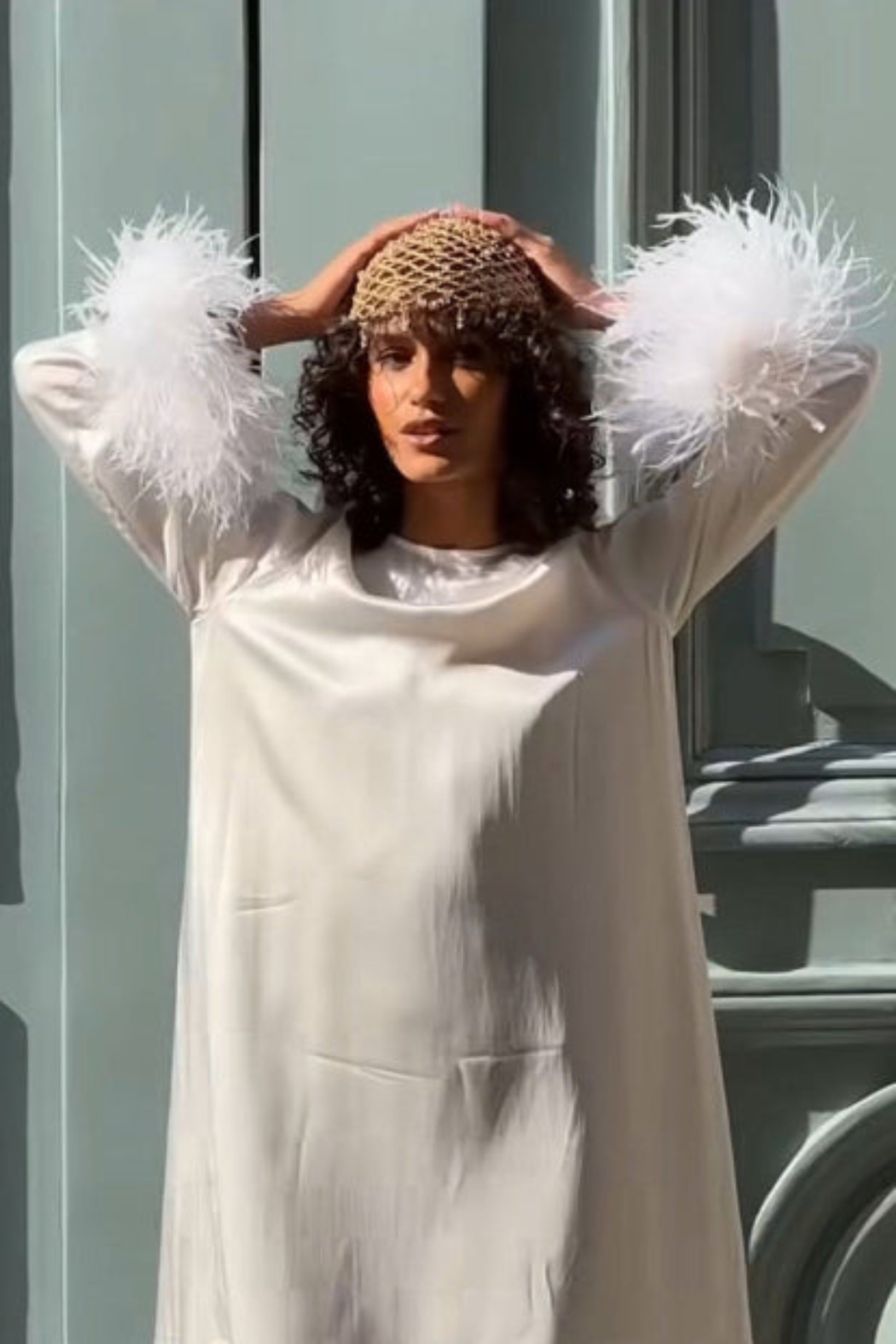 Woman wearing a gold crocheted and glass crystal beaded hat in a white dress with feathered sleeves standing in front of a green door.