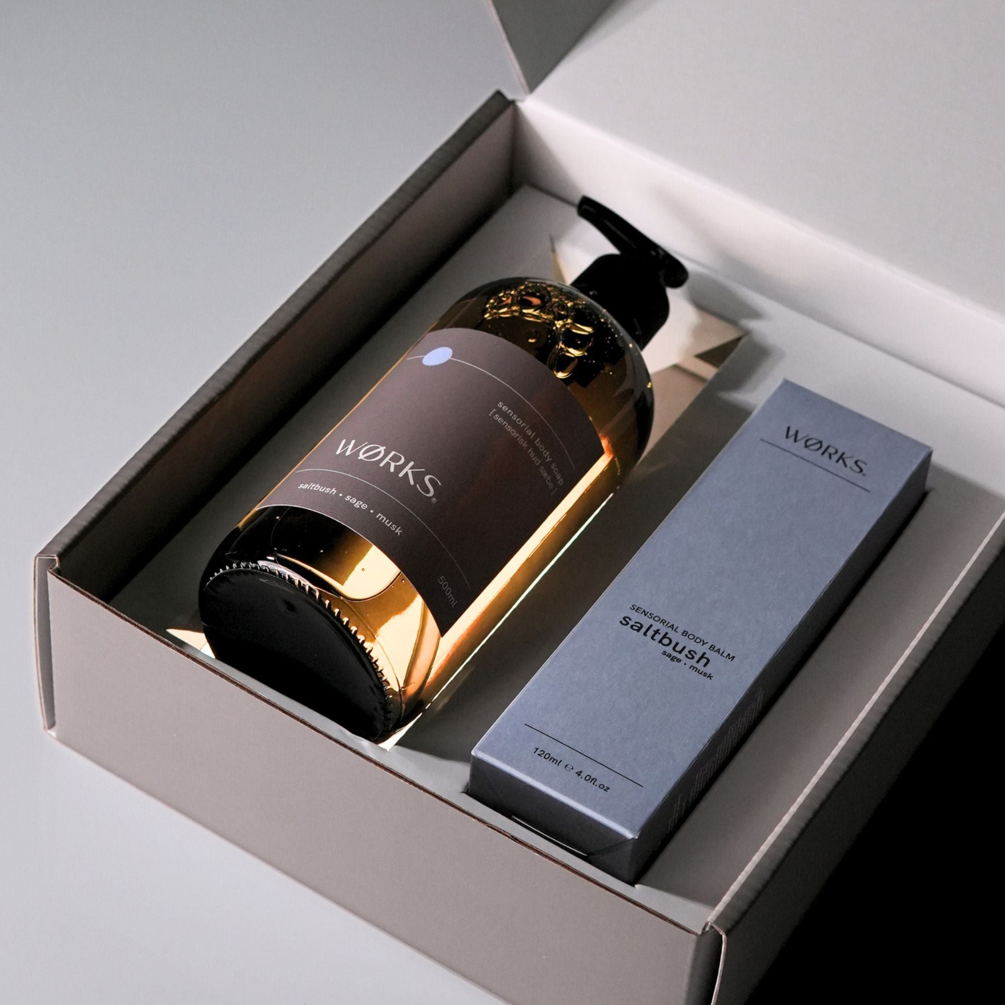 A brown, glass pump bottle and a blue box of 'WORKS' products in a grey gift box with lid open.