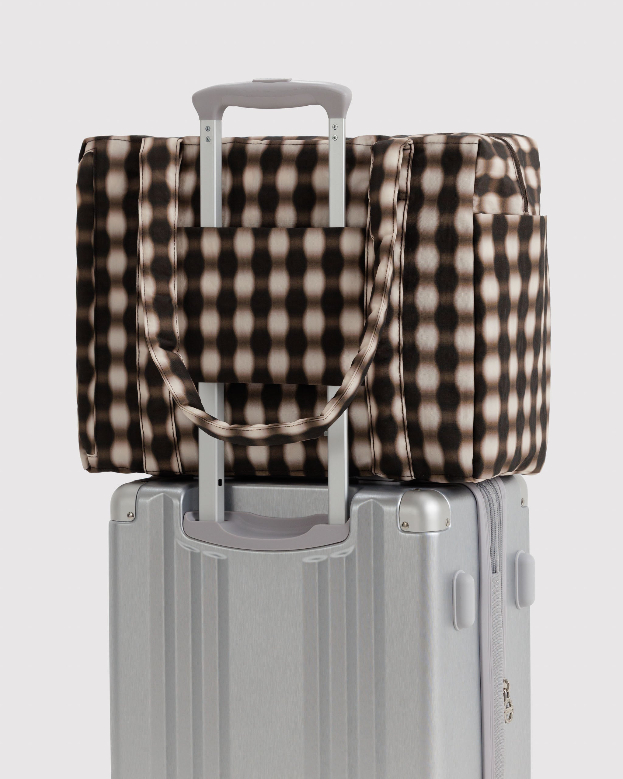 Checkered blurry brown and ecru tote bag on top of a silver suitcase attached by the luggage sleeve at the back of the bag.