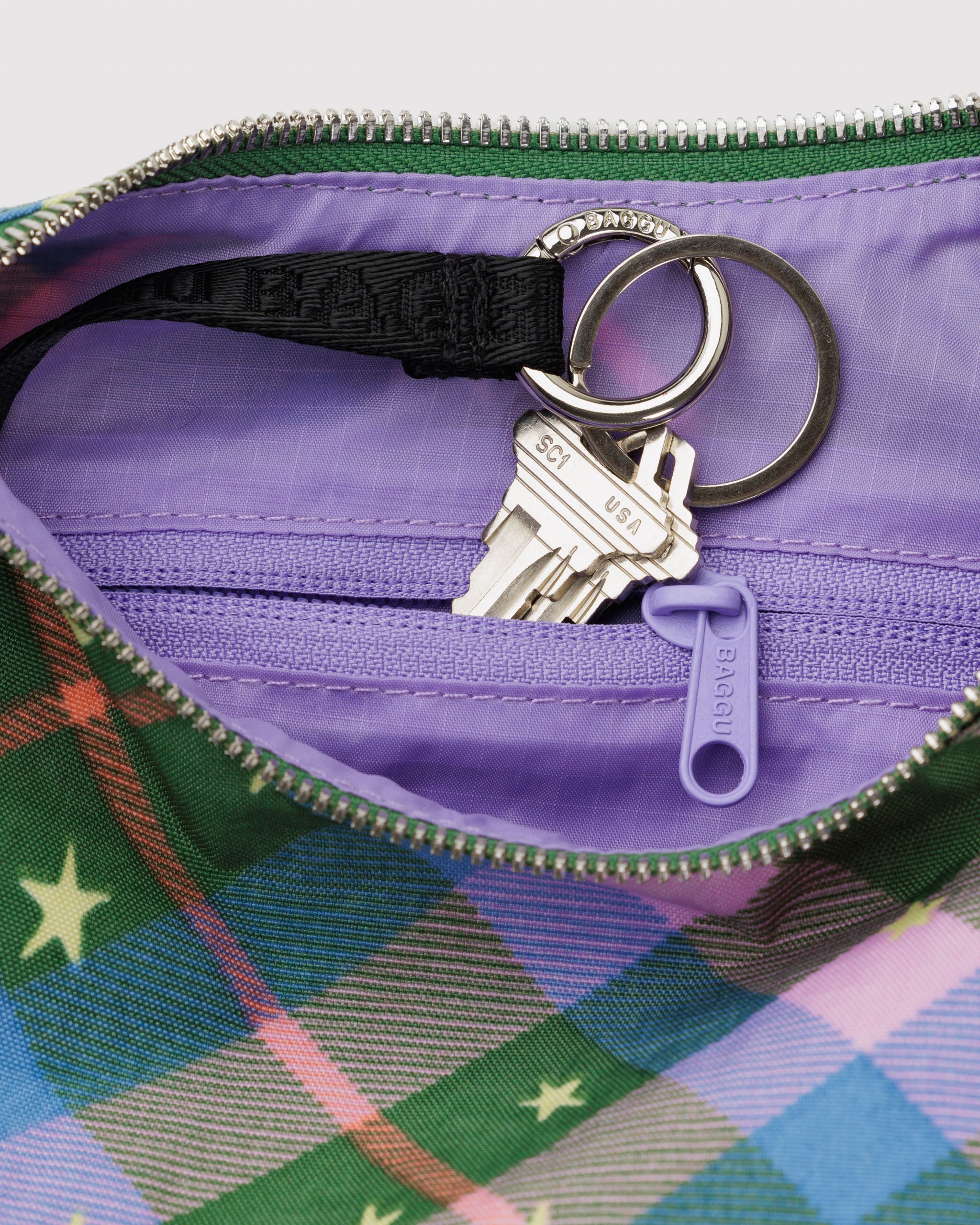 Close-up of a purple lining, zip pouch and black key leash with keys inside green plaid bag with stars.