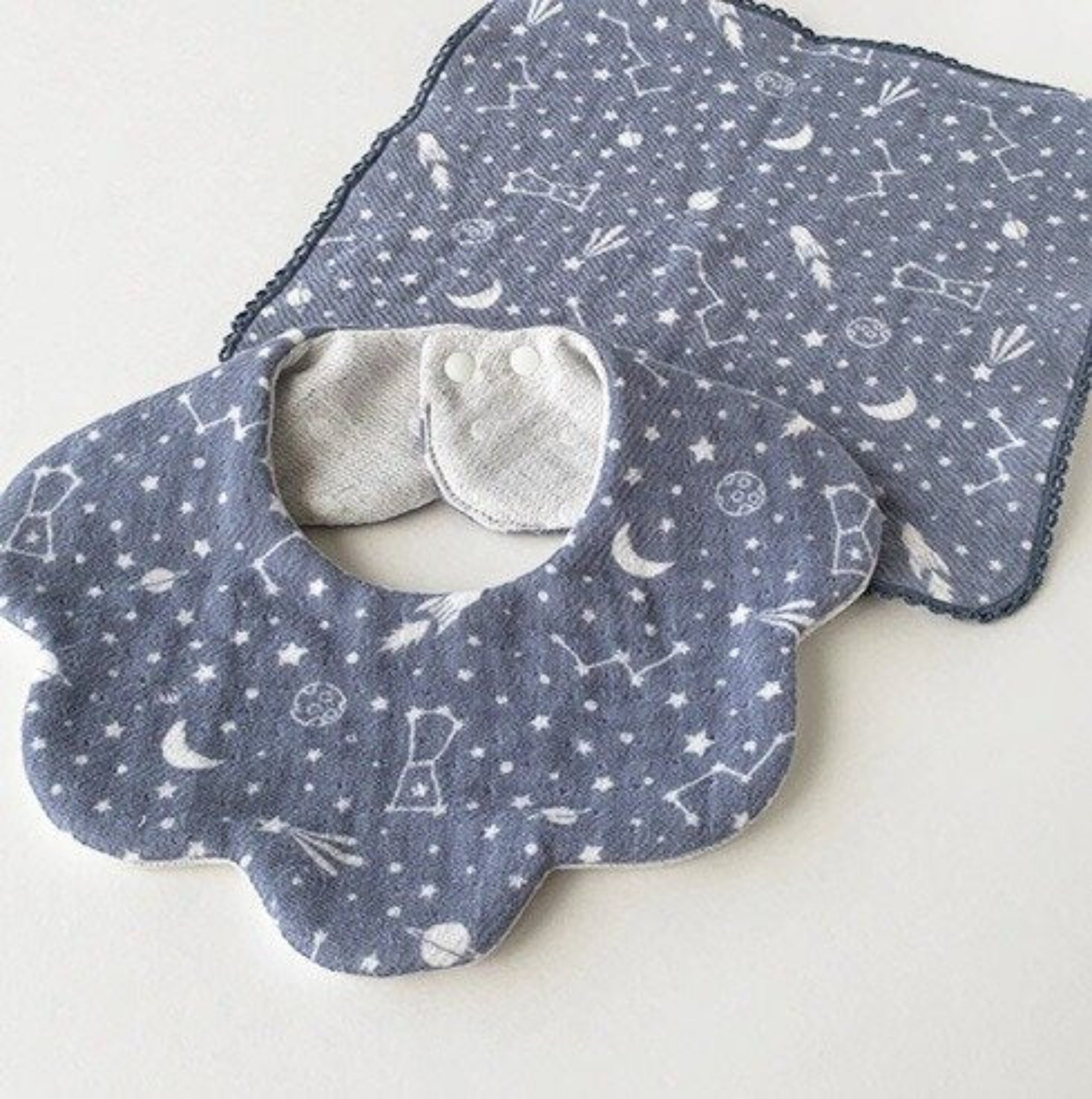 Blue grey space-patterned baby bib with white dot jacquard reverse side in a scallop edge design and two snap closures lying on top of a face towel in same pattern.