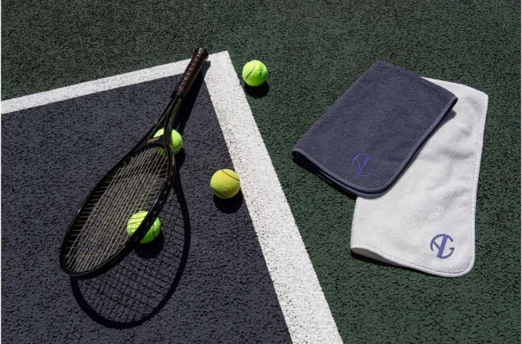 White and a blue sports towel with AG logo folded on tennis court next to racquet and tennis balls.