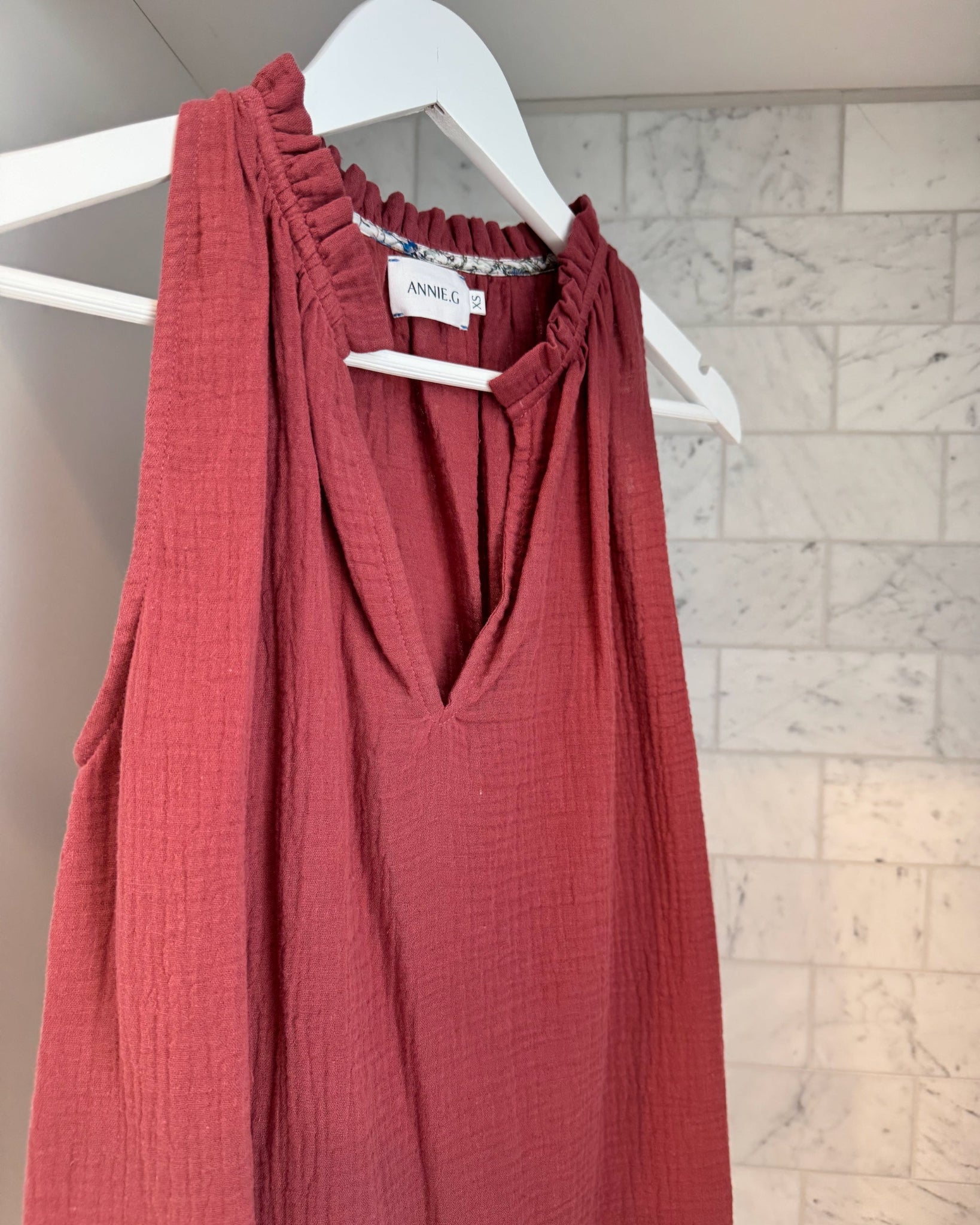 Close up of a maroon sleeveless top with v-neckline split and frill with floral bias binding on inside neck on a hanger.