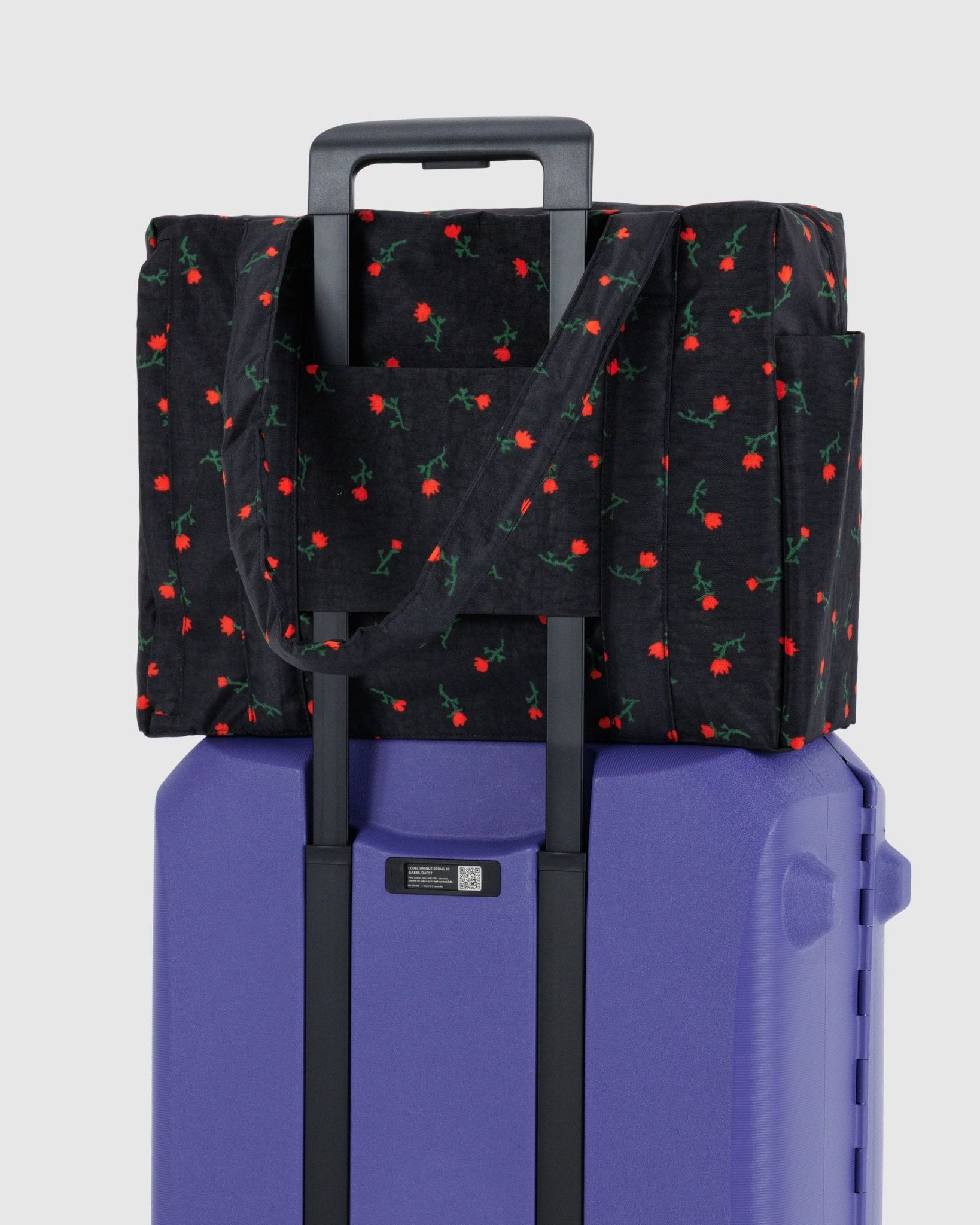 Small, black duffle bag with red wildflowers on top of a purple suitcase attached with travel sleeve at the back