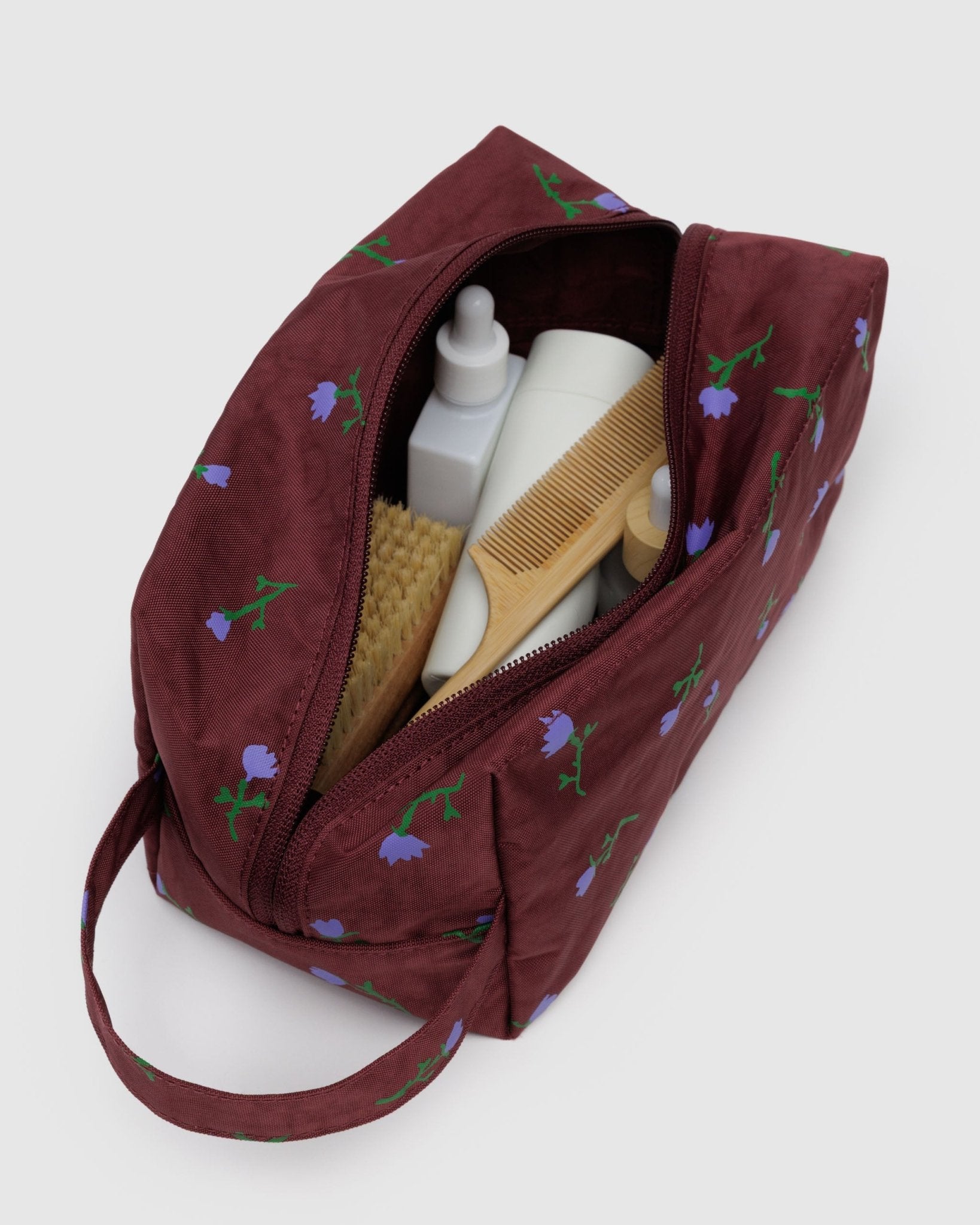 Maroon toiletry bag with purple wildflower patterns and carry handle containing a bottle, brush, and comb.