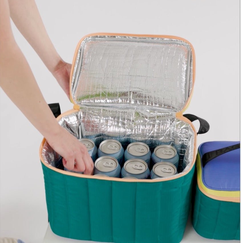 A persons arm reaching into an open zip insulated bag with drink cans sitting next to a closed puffy lunch bag in same colour.