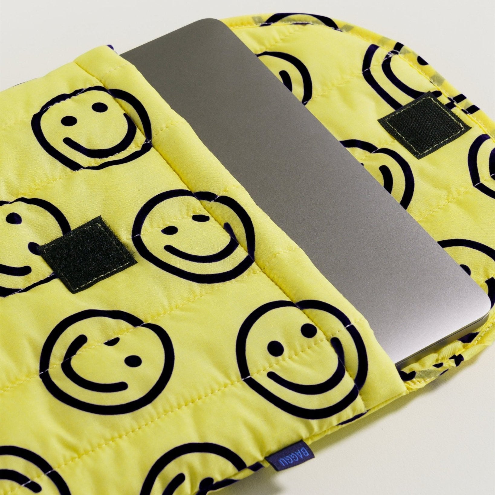 Yellow quilted laptop sleeve with smiley face print in black outline with open velcro closure top and silver laptop inside.