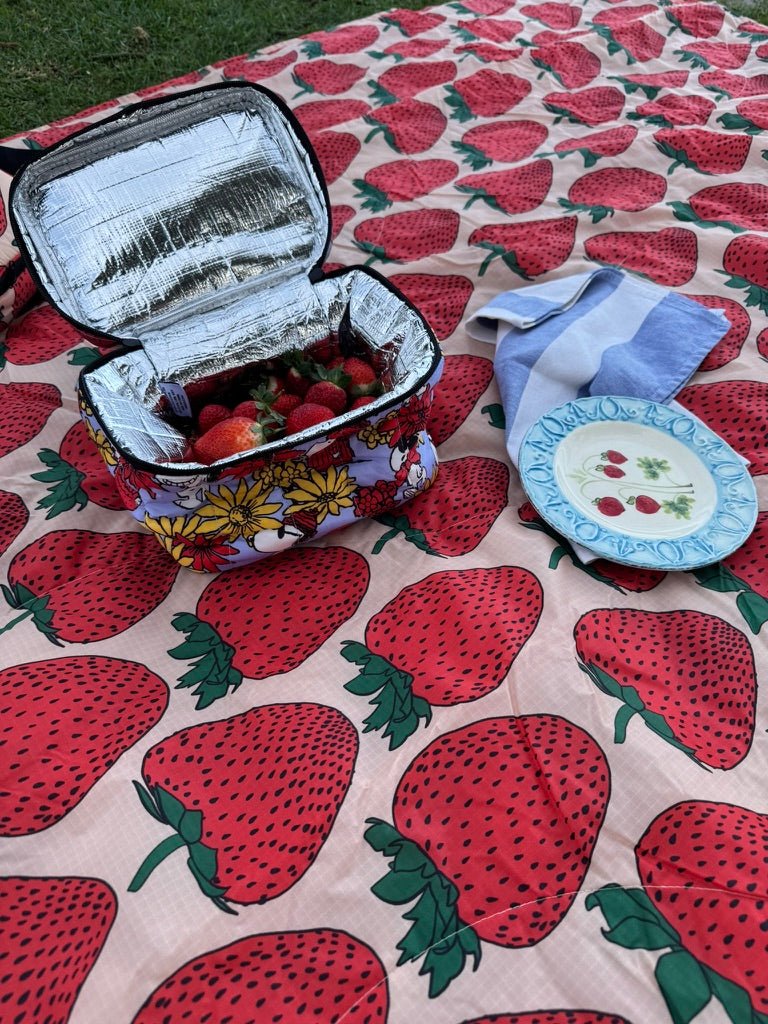 Insulated puffy lunch bag with strawberries inside, a plate and blue and white wide striped tea towel on a strawberry-patterned quilted picnic blanket.