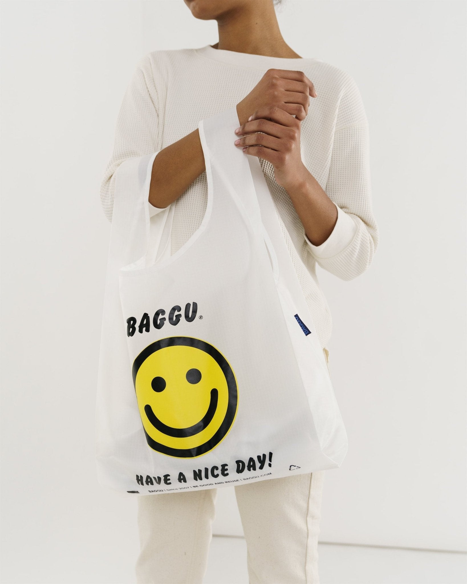 Person holding a yellow smiley face Standard Baggu Reusable Bag on arm across body