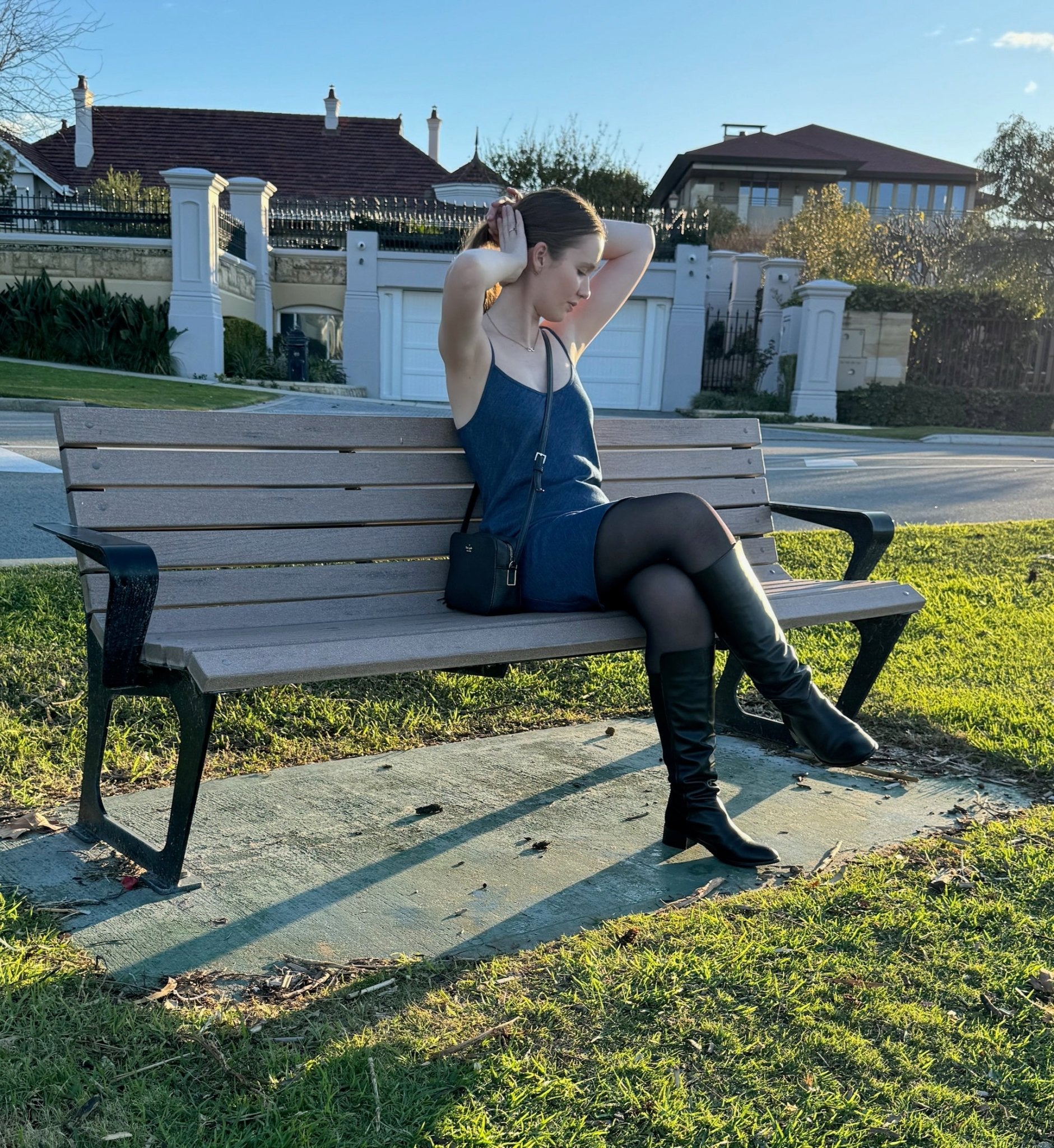 Woman wearing blue denim Bias Slip Mini Dress and black boots and bag sitting on a bench in a park.