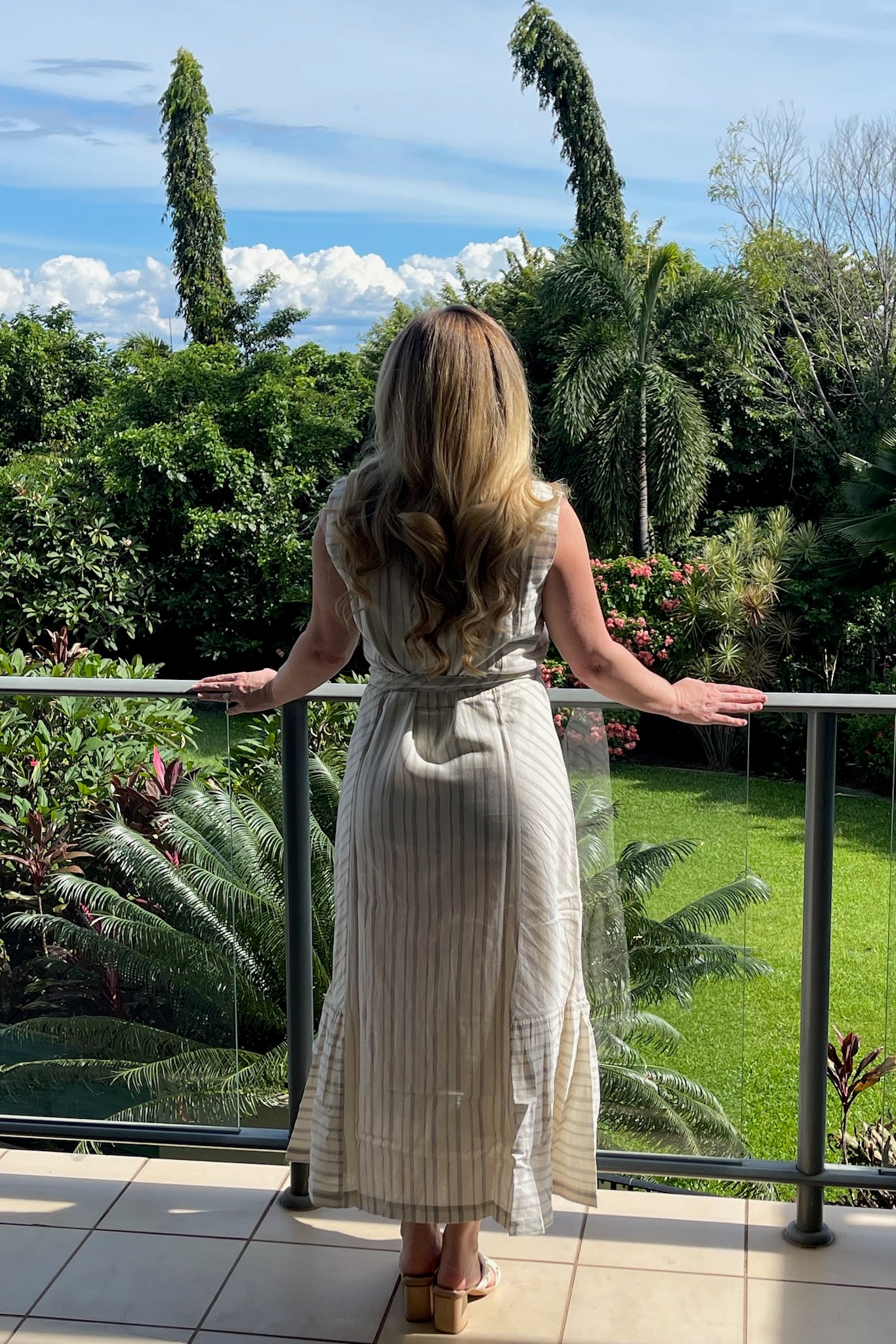 woman-wearing-cream-and-indigo-striped-sleeveless-dress-with-belt-standing-on-balcony-back-view