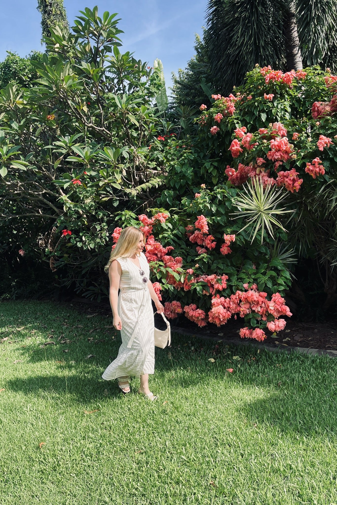 woman-wearing-cream-and-indigo-striped-sleeveless-dress-with-belt-walking-on-grass-near-pink-flowers-holding-bag-with-sunglasses-tucked into-v-neckline