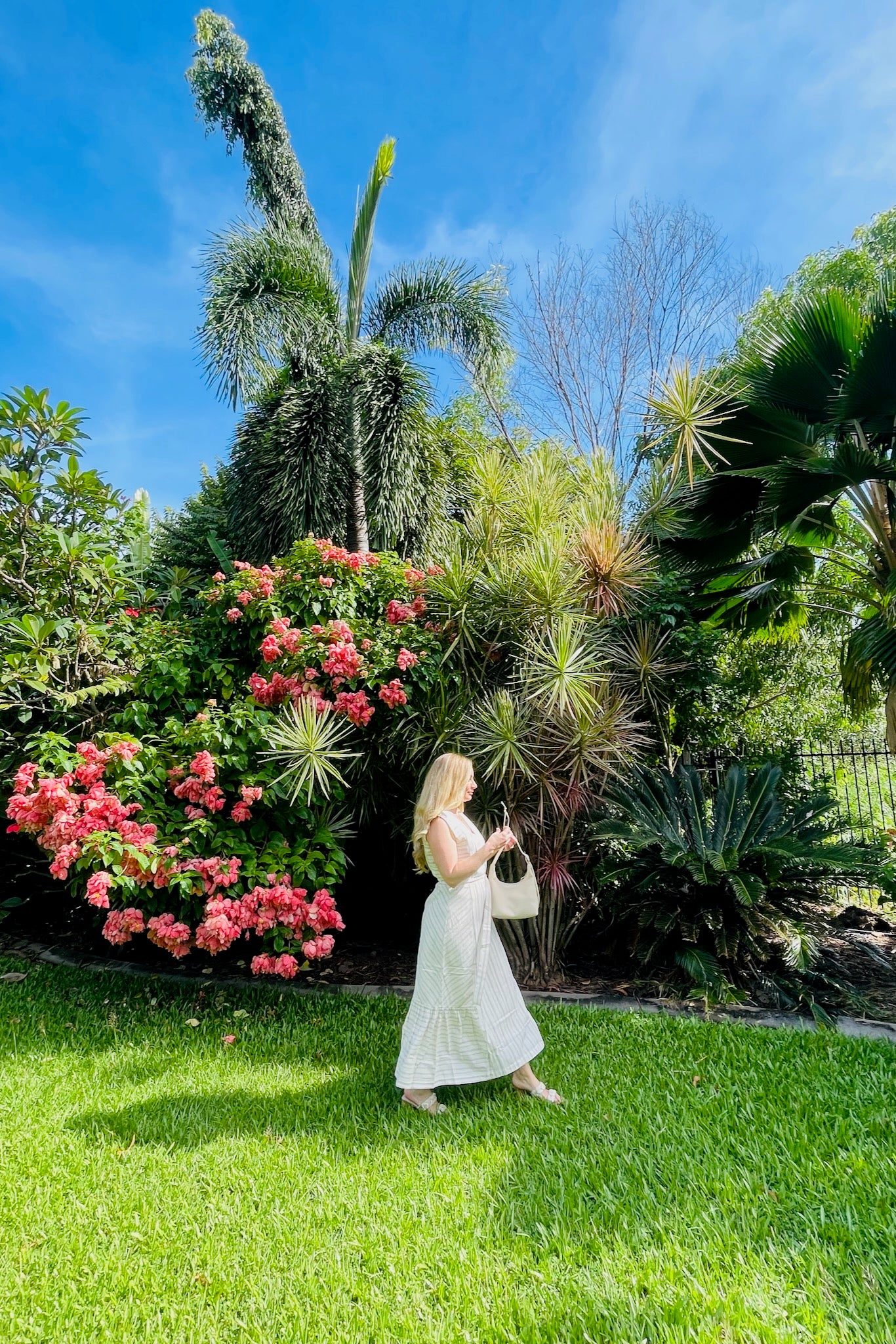 woman-wearing-cream-and-indigo-striped-sleeveless-dress-with-belt-holding-a-bag-walking-in-garden-side-view