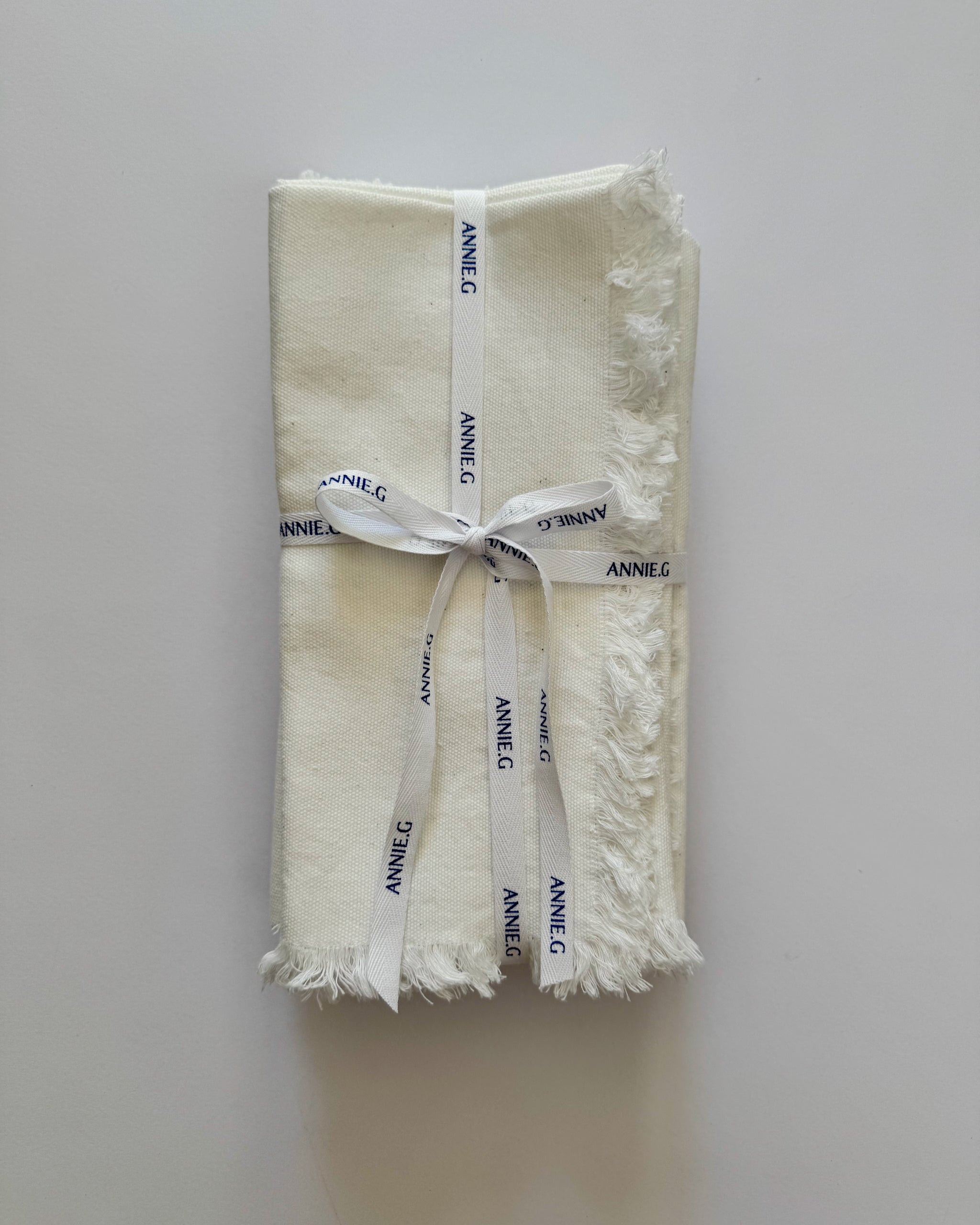 Folded and stacked frayed edge white cotton napkins with 'ANNE G' branding ribbon tied around.