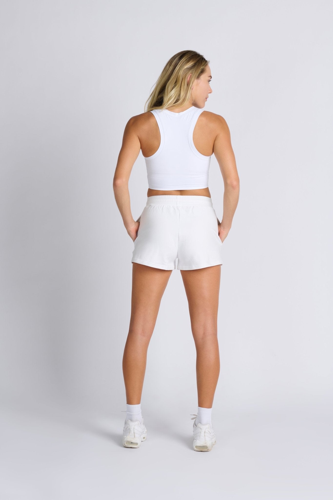 Back of a woman wear white Monterey Sweat Shorts and racer back crop top with hands in pockets.