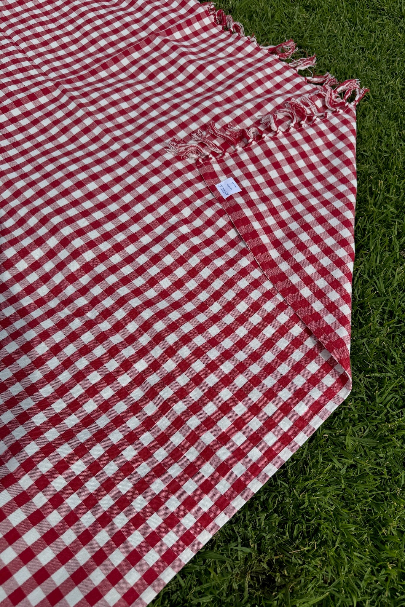 Red and white checked Gingham Cotton Tablecloth on green grass as a picnic blanket showing brand label - ANNIE.G