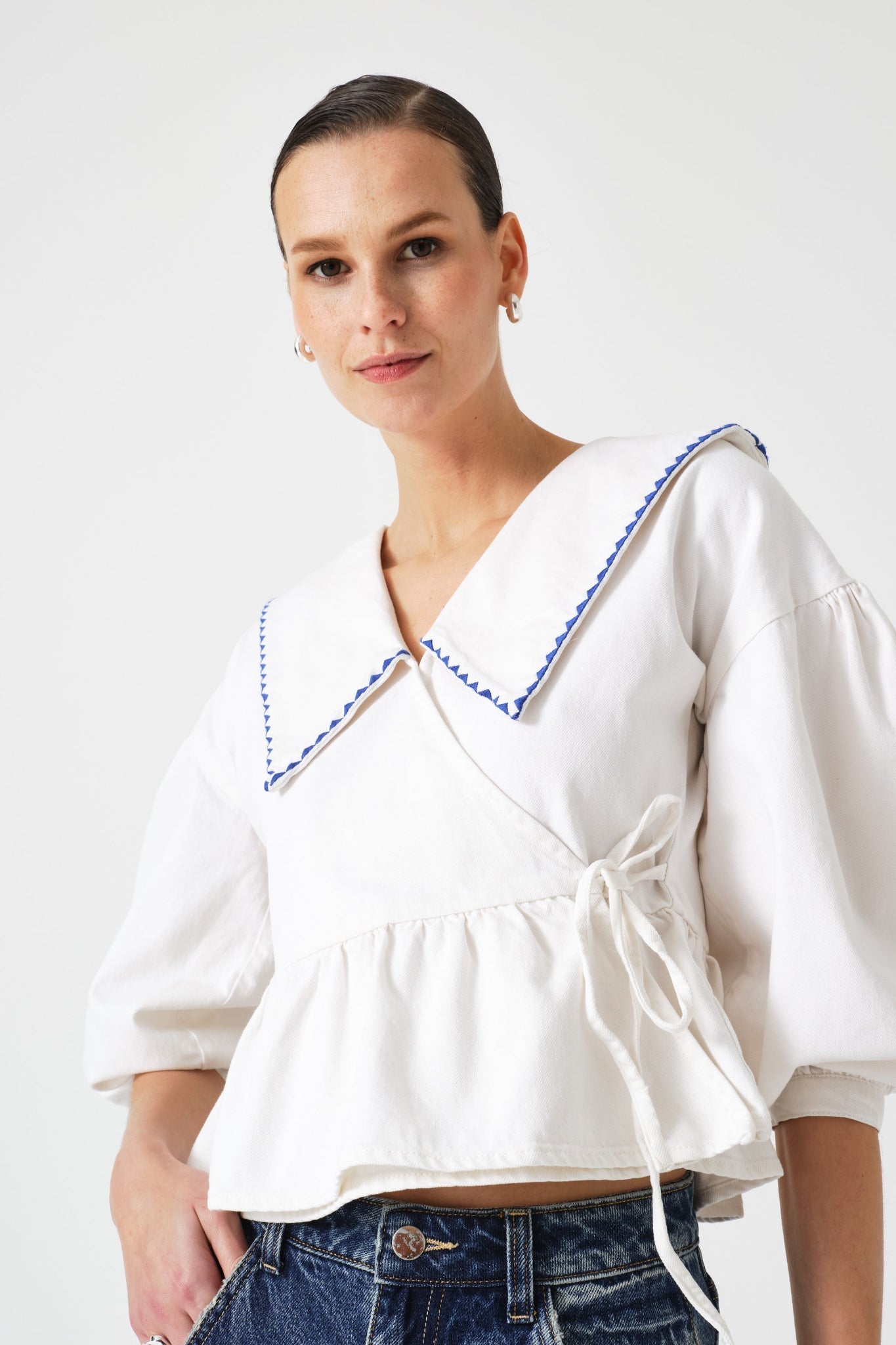 Woman wearing a white wrap blouse with a blue trim on collar and tie at side with peplum hem