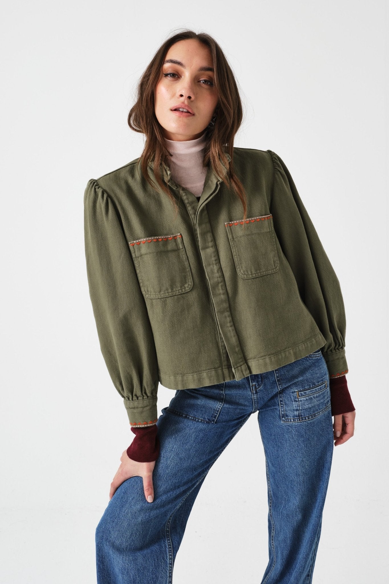 Woman wearing an olive green jacket and blue jeans leaning arm on bent leg