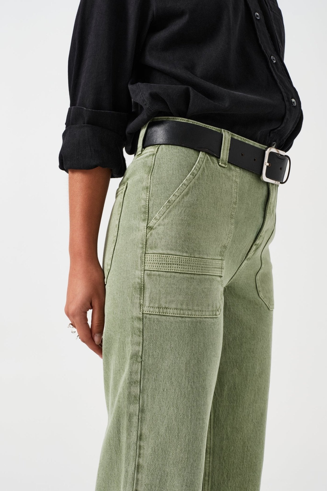 Close up of the side of a person wearing green jeans with retro patch pockets and a black belt through loops with a black shirt.