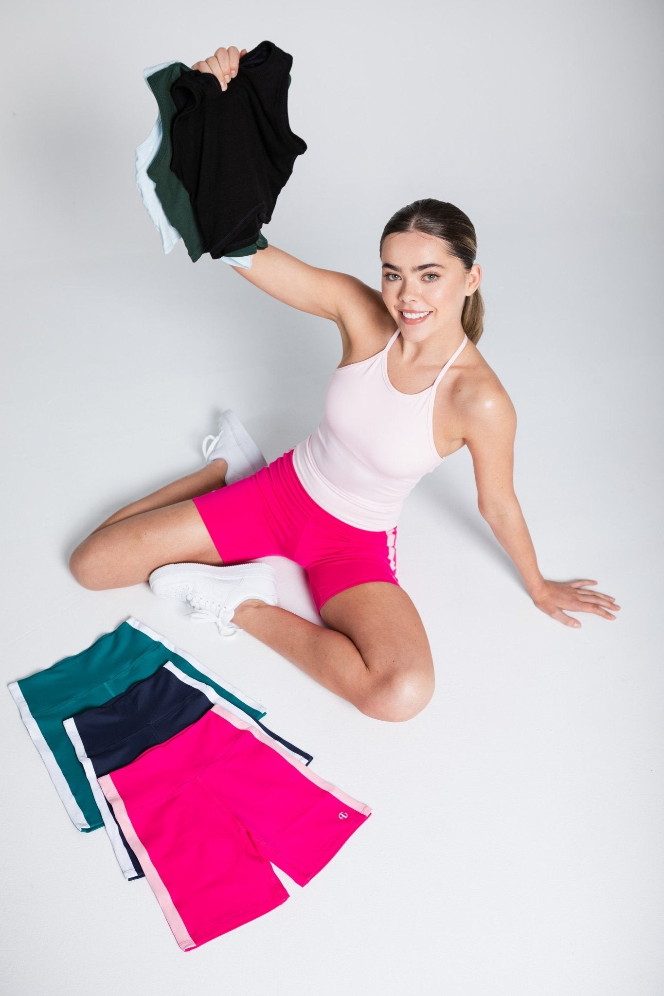 Woman in pink activewear top and shorts sitting surrounded by bike shorts and holding crop tops up with one hand.