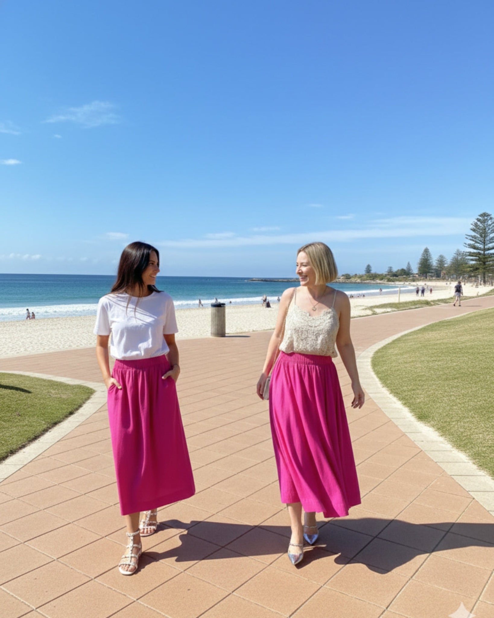 Two women walking on a boardwalk by the beach with clear blue skies wearing a hot pink elastic waist skirt and different tops.