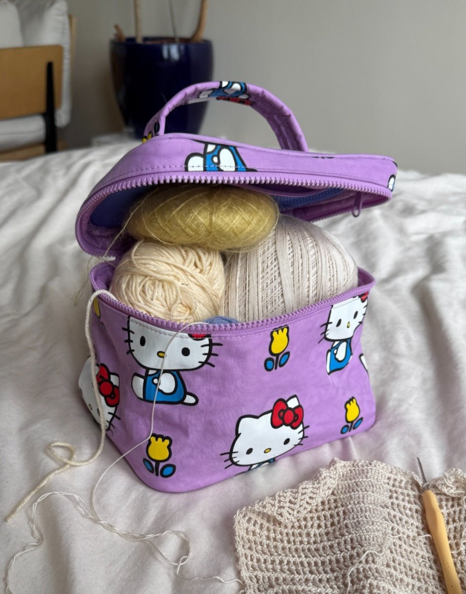 Purple Hello Kitty-patterned cosmetic case used for storing yarn inside for knitting project on a bed
