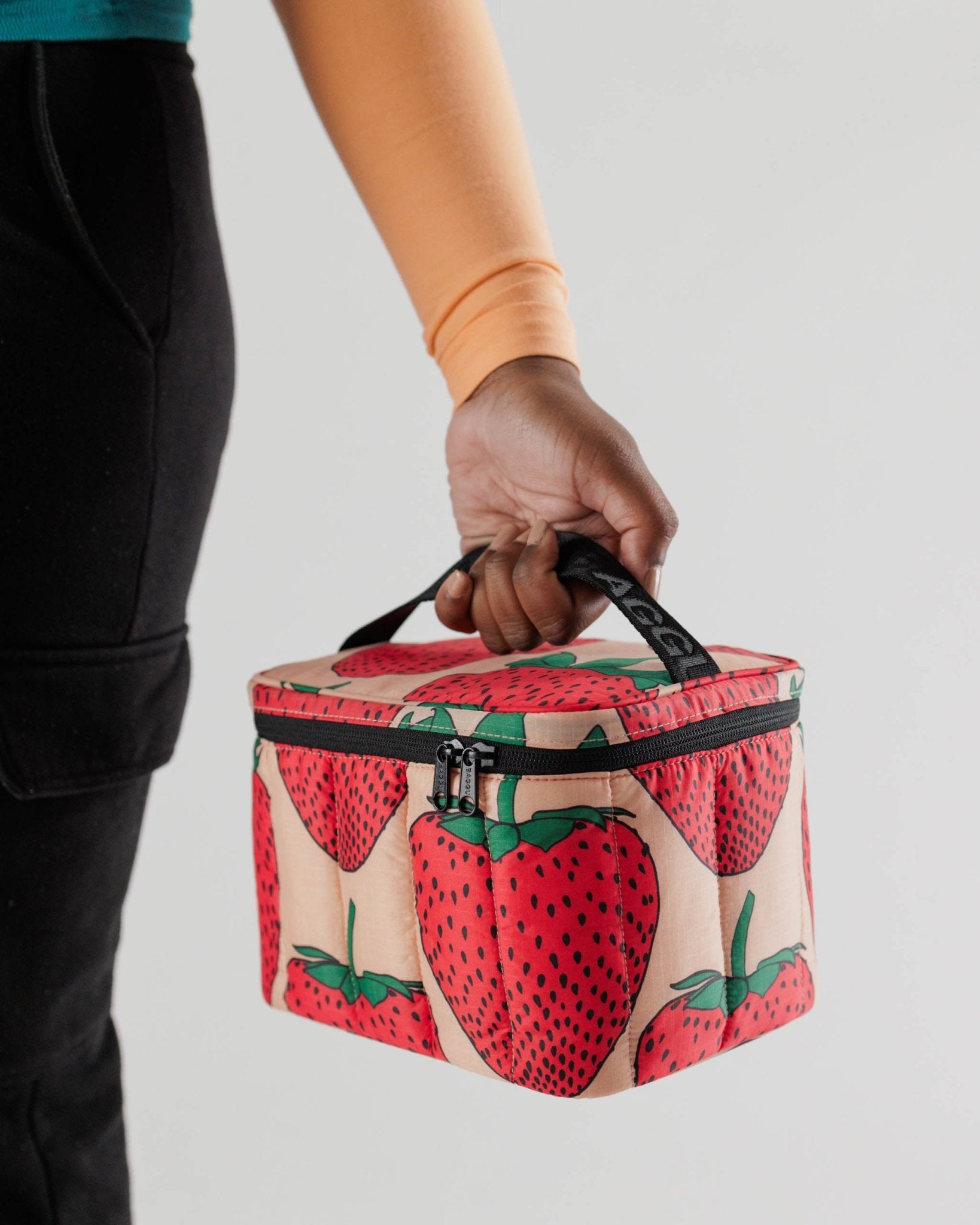 Person holding the black top handle on a strawberry print Puffy Lunch Bag with a black zip.
