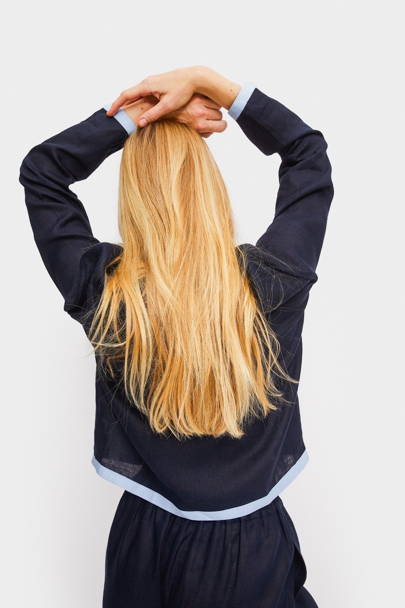 Back of a woman with hands on head wearing a navy embroidered shirt with long sleeves and contrast light blue trim with matching long pants.
