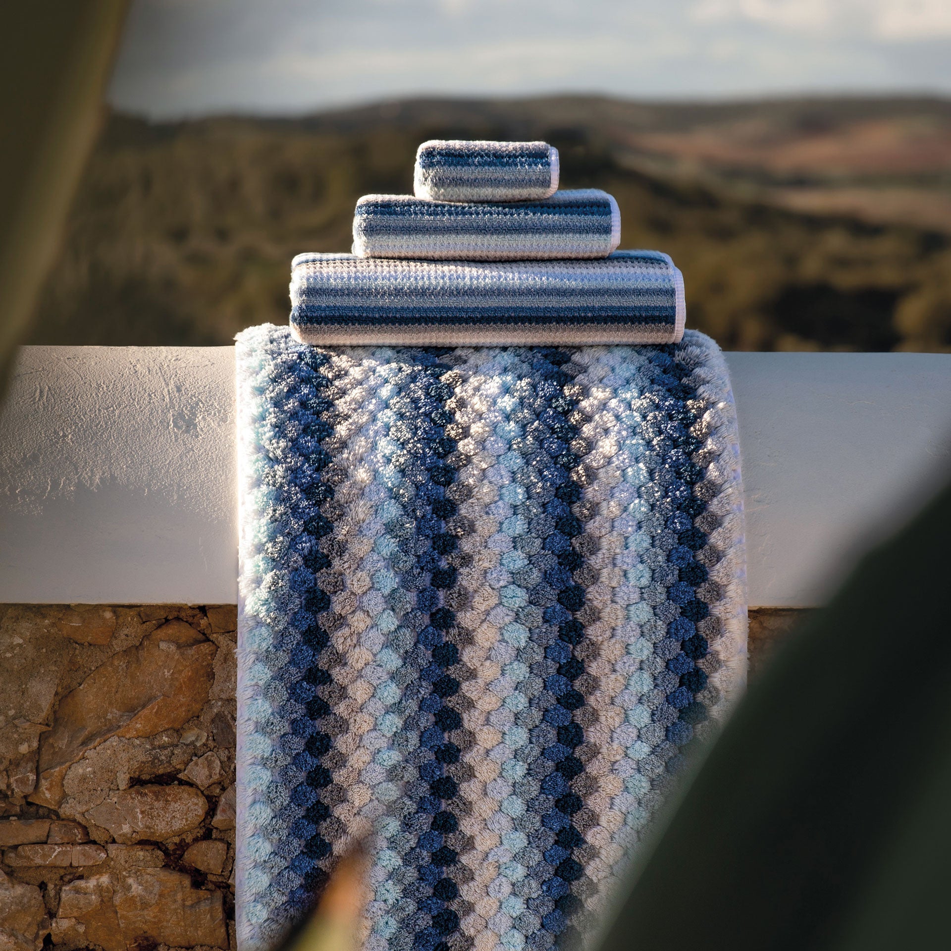 Blue multi stripe bath rug mat and with a stack of folded matching towels on top outside in landscape.