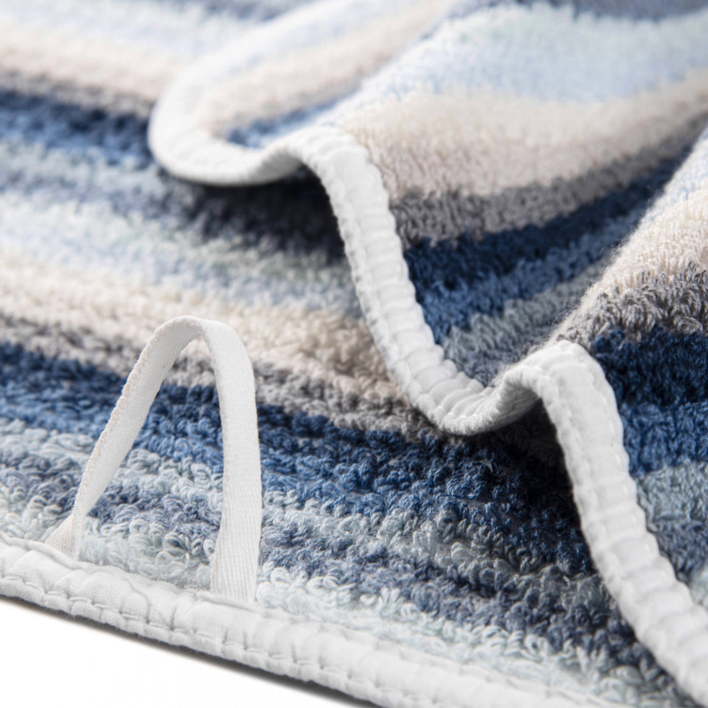 Close-up of towels textured blue multi stripe cotton print with a white hanging loop and white bias binding edge.