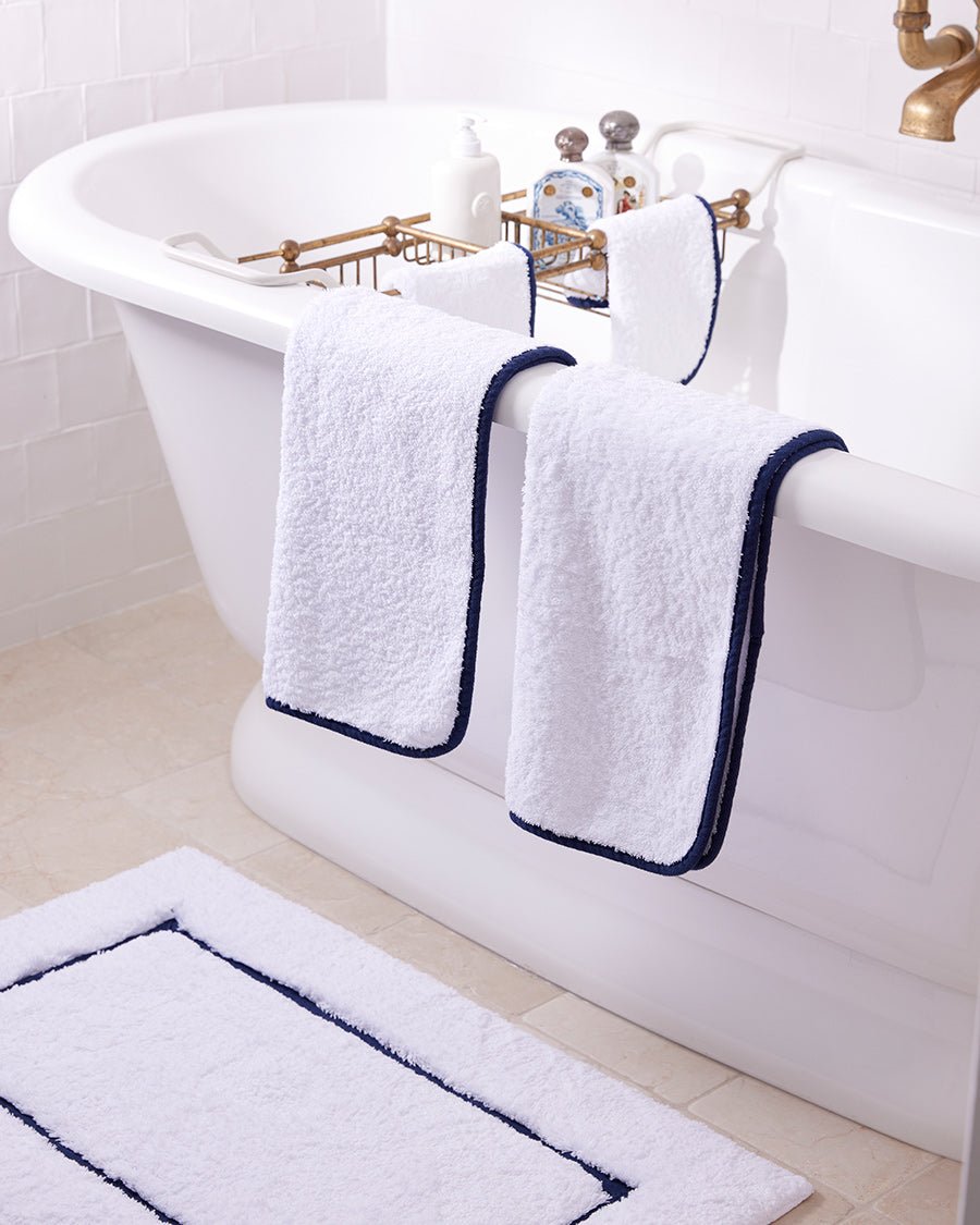 Folded white bath towel with navy bias binding edge on bath tub edge and matching bath mat on the floor.